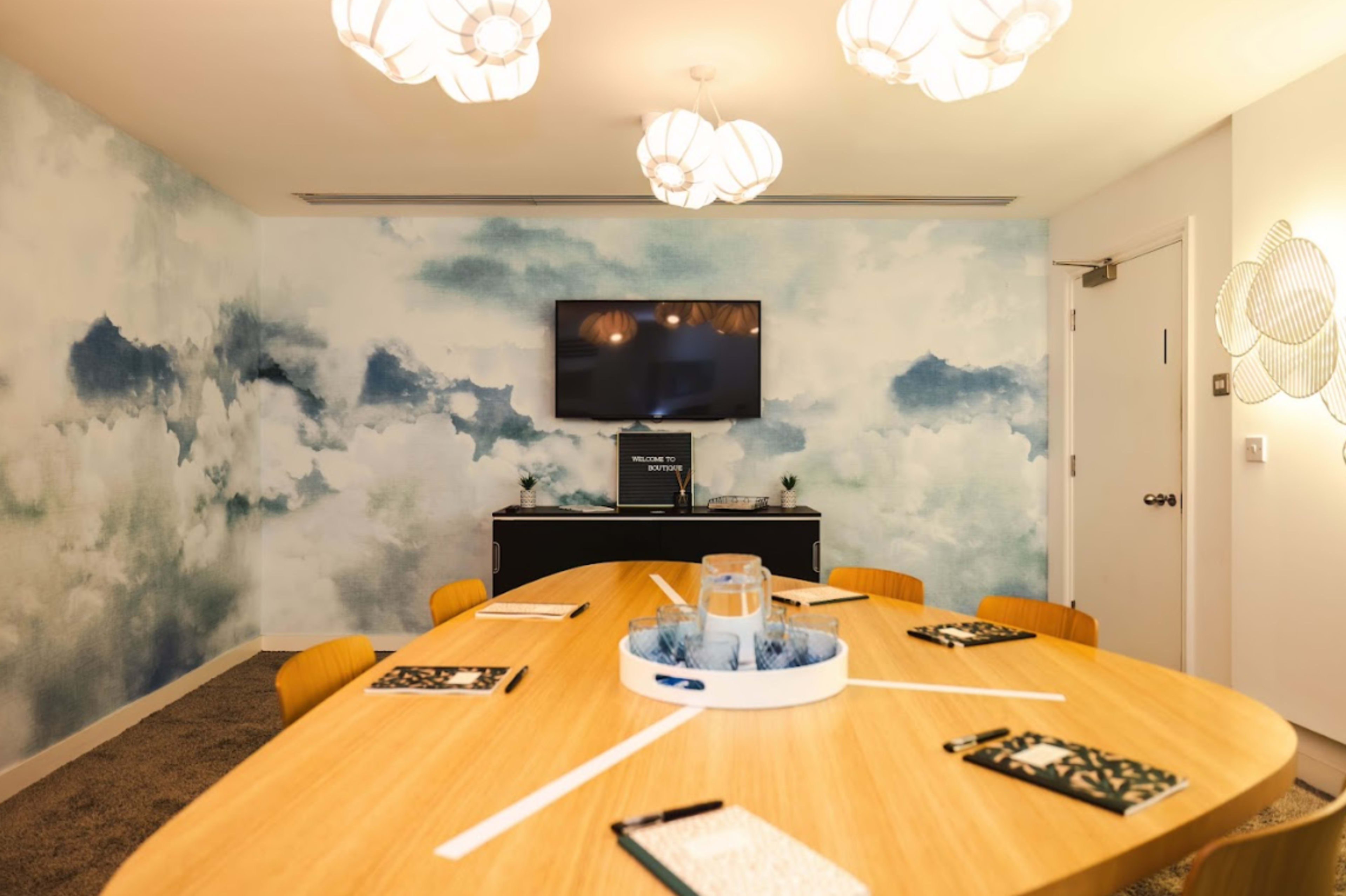A modern conference room with a large wooden table, several notebooks and pens arranged around it, and a wall adorned with a blue cloud-patterned mural.