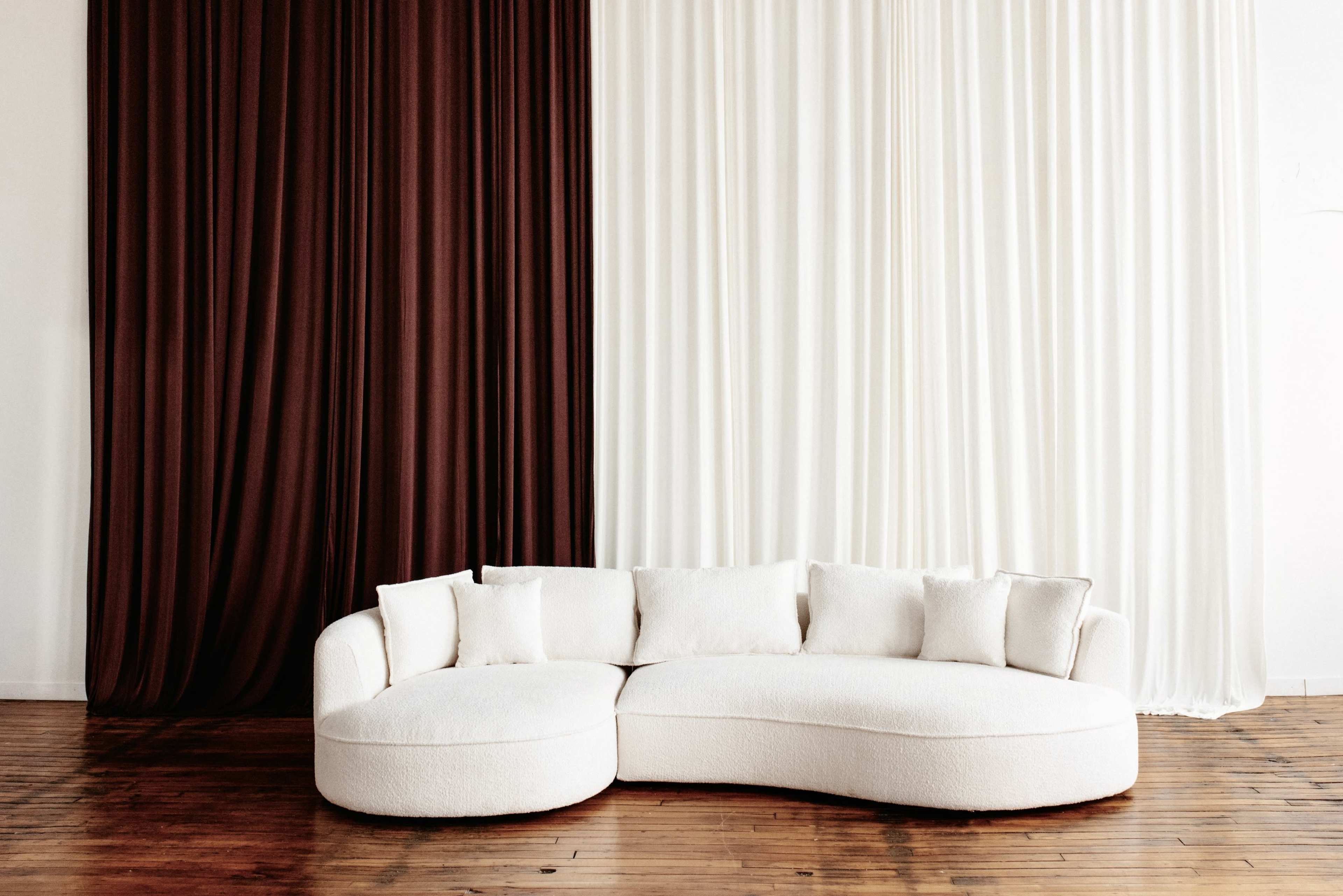 The image features a curved, cream-colored couch with multiple cushions placed against a backdrop of contrasting brown and white draped fabric.