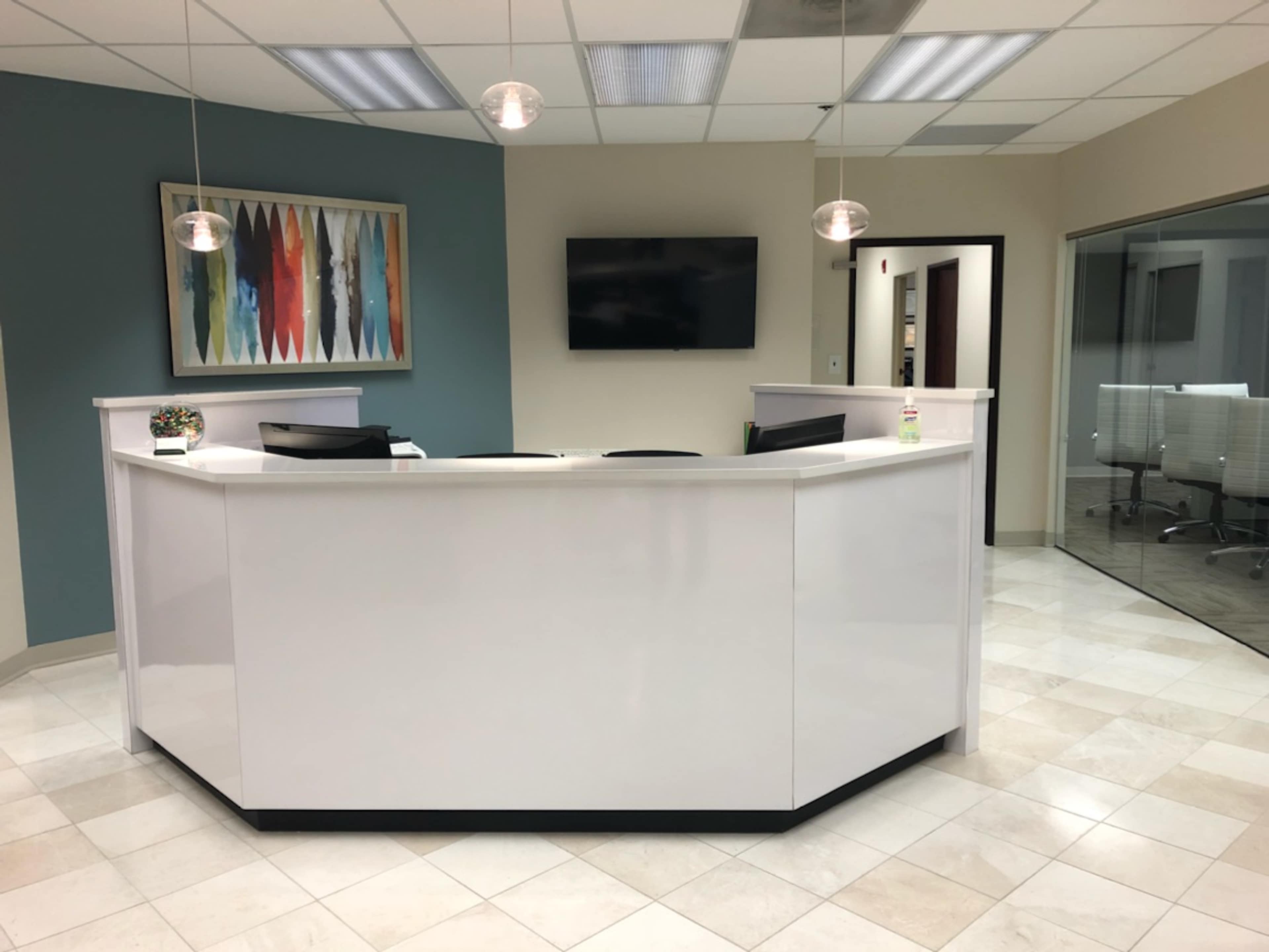 A modern reception area featuring a white front desk, pendant lights, a wall-mounted television, and a colorful artwork in the background.