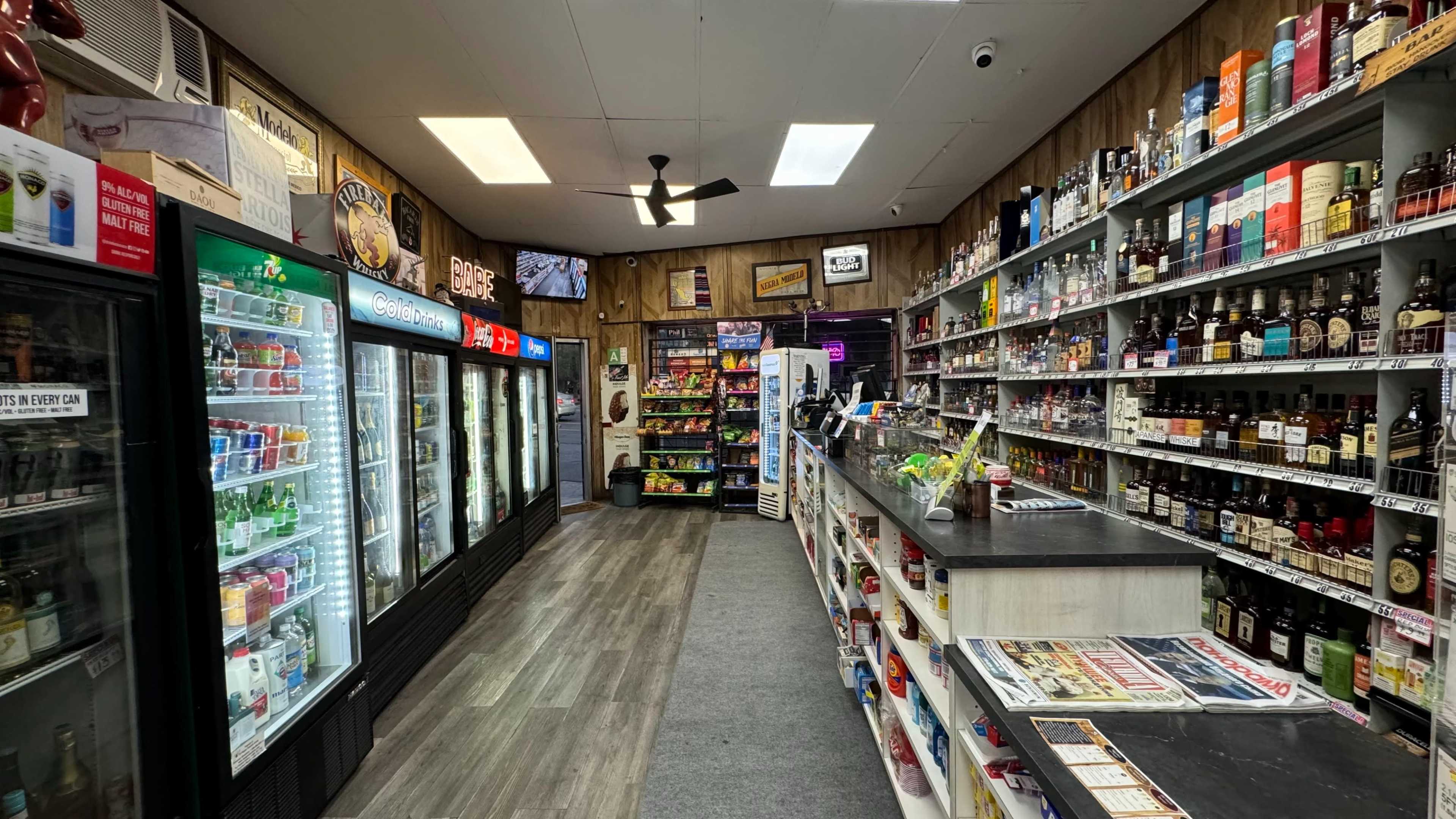 The image shows the interior of a convenience store featuring shelves stocked with various beverages and snacks, a checkout counter, and refrigerators displaying drinks.