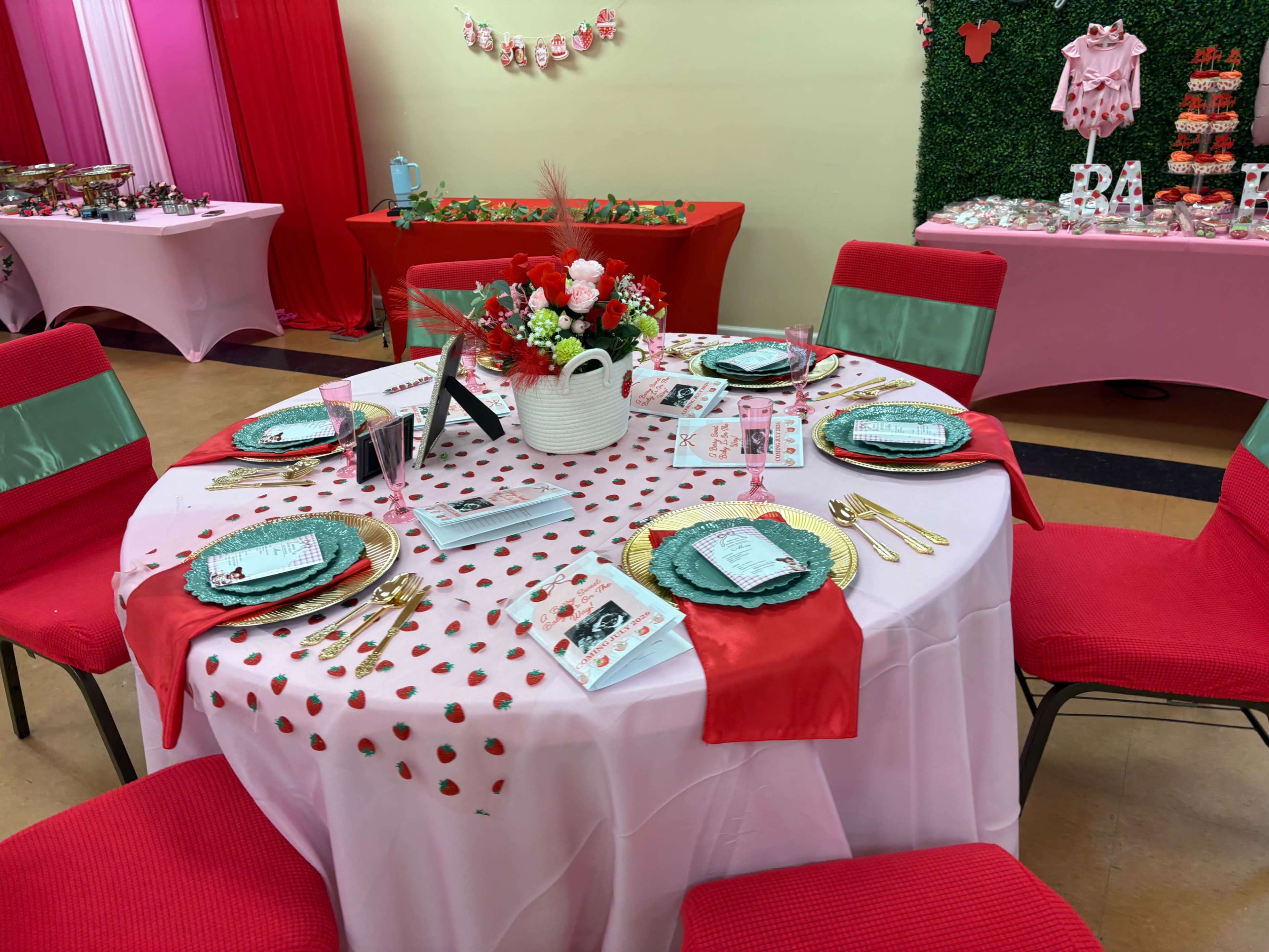 A round table is elegantly set with green and red plates, floral decorations, and menu cards, surrounded by red chairs in a festive event space.