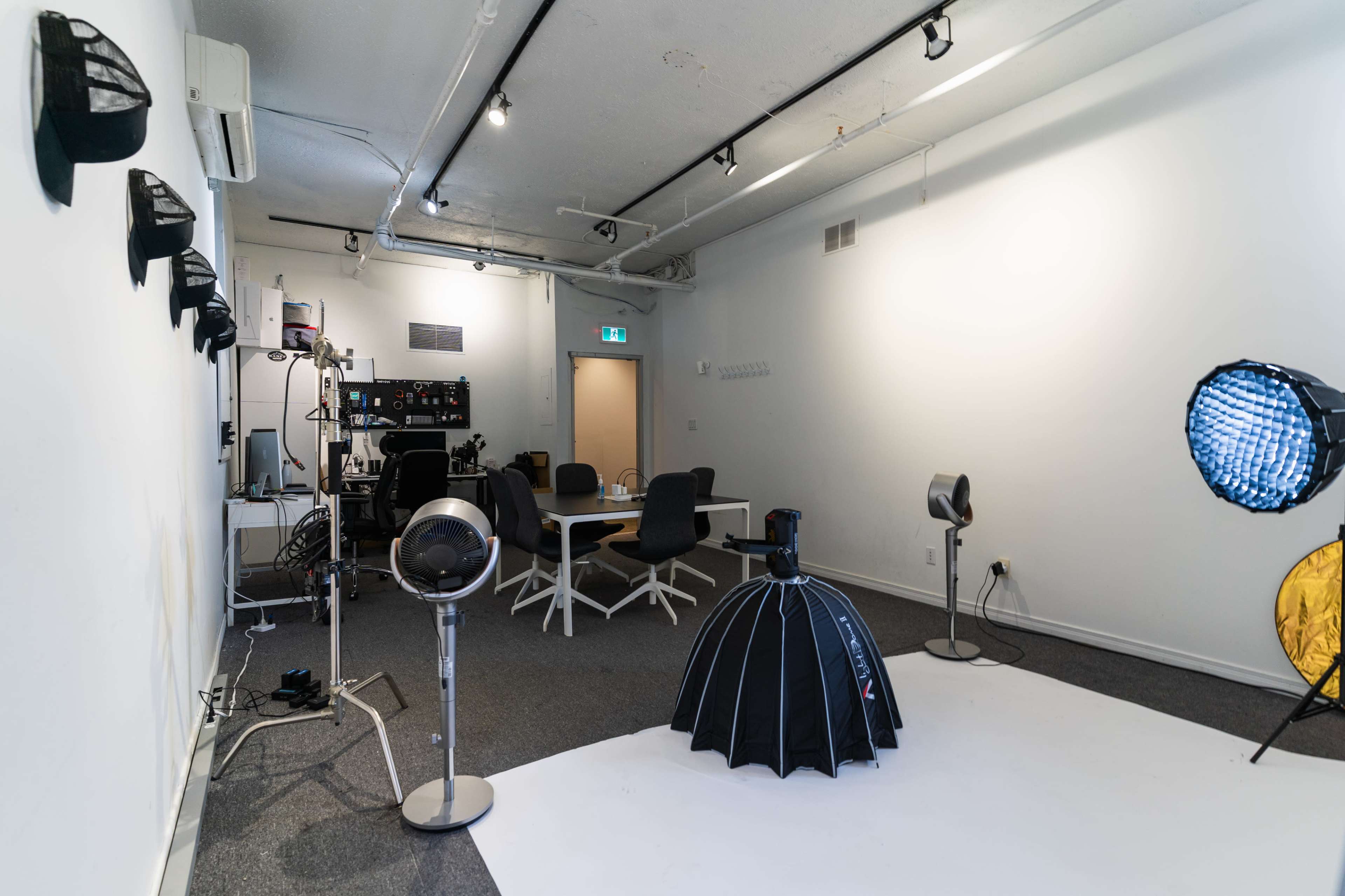 The image shows a photography studio featuring various lighting equipment, a white backdrop, a table with chairs, and a control station against the wall.