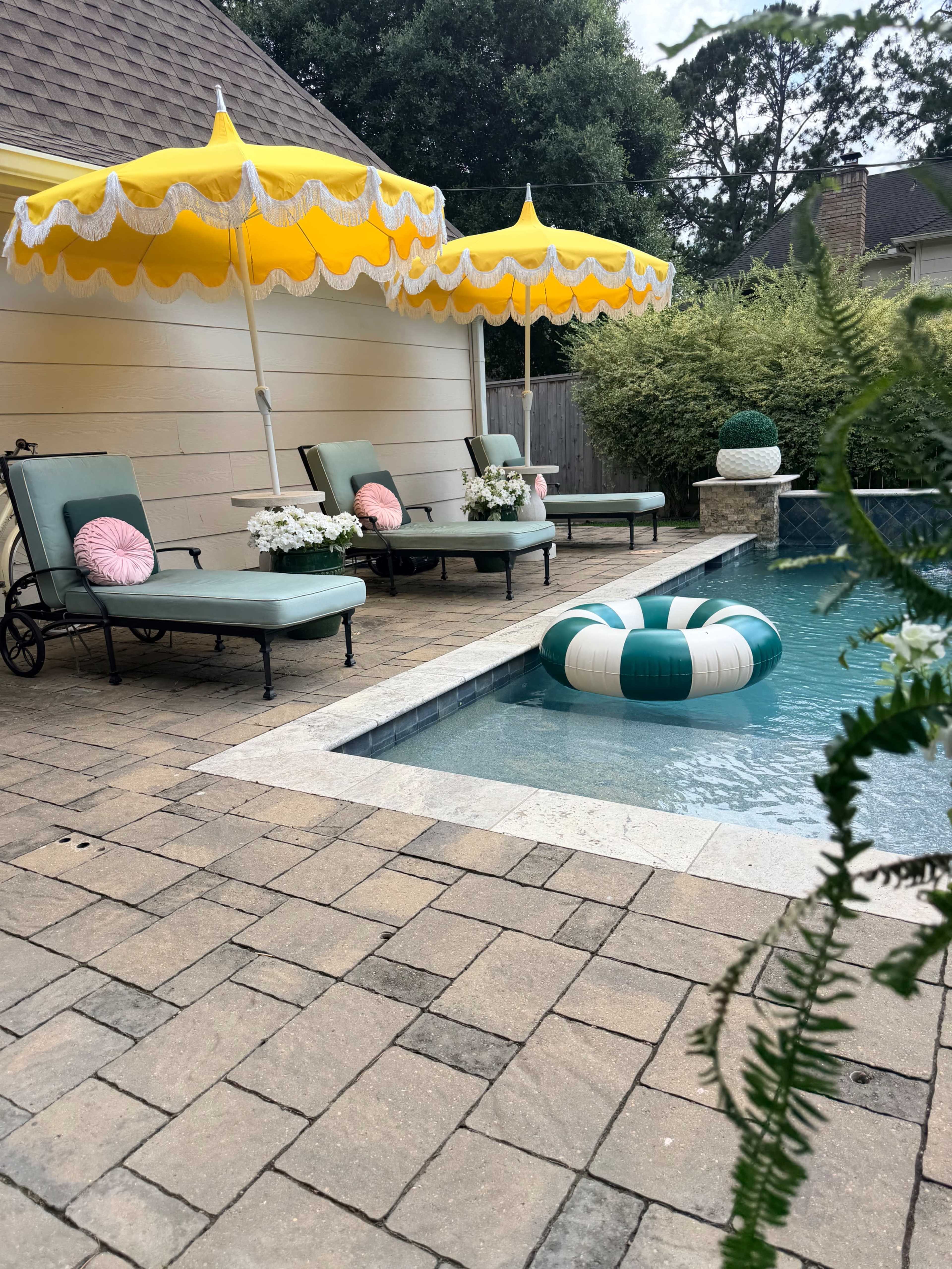 The image shows a backyard pool area with two lounge chairs, yellow umbrellas, and a large inflatable ring floating in the water.