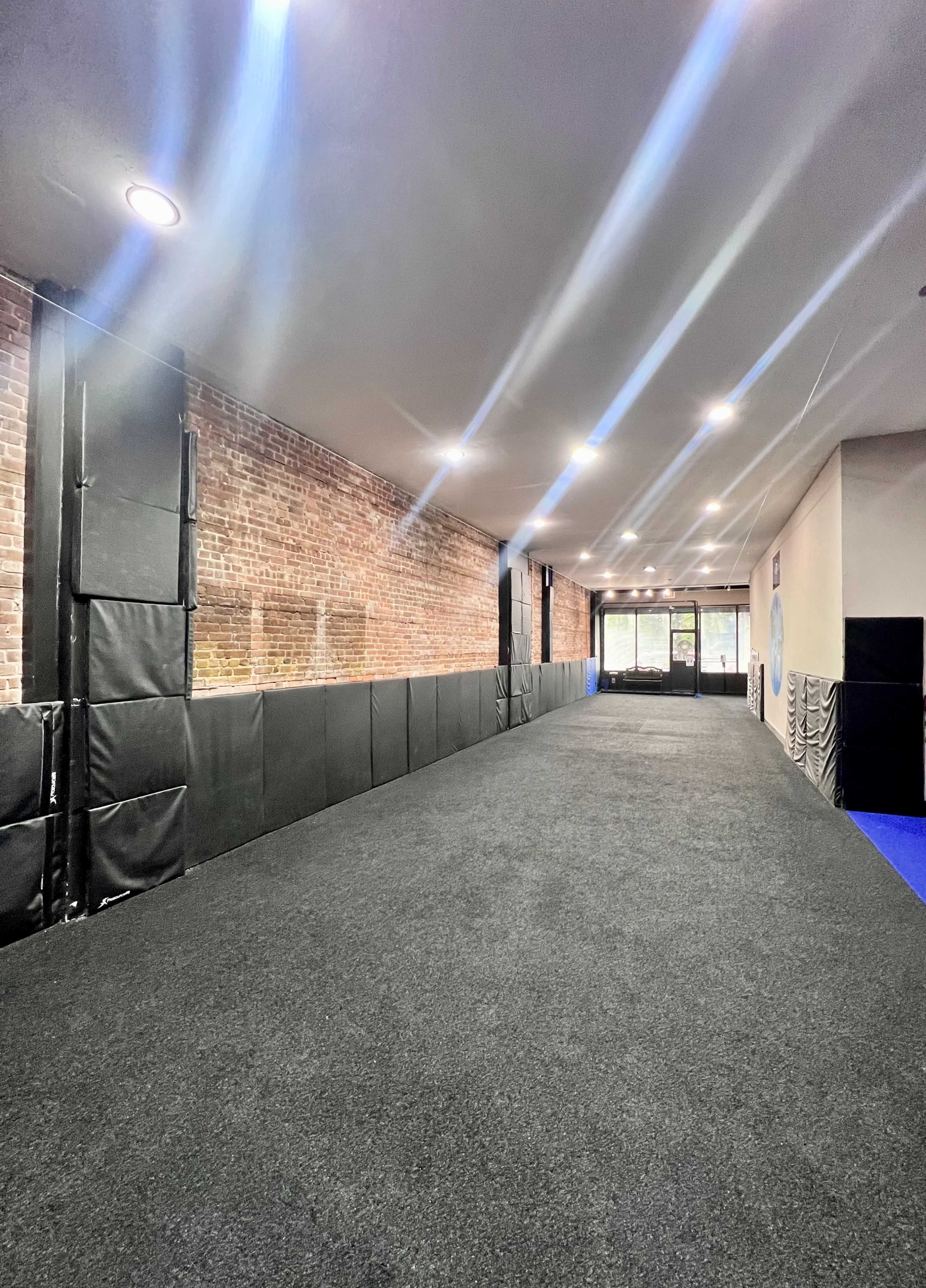 A long, narrow hallway with a dark carpeted floor, brick walls, and padded black wall protection, leading to glass doors at one end.