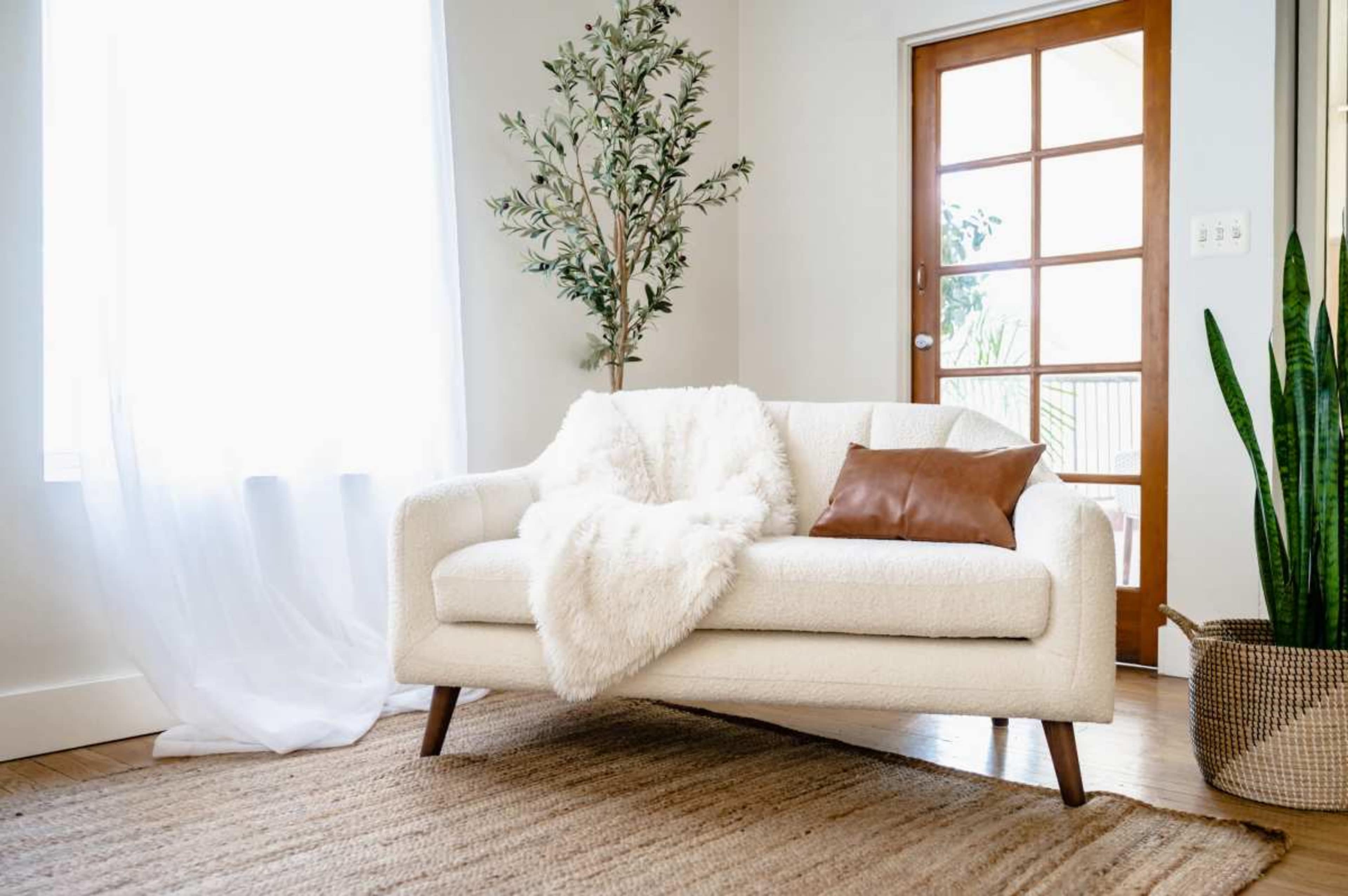 A light-colored sofa with a throw blanket and a leather cushion is positioned beside a door with glass panels, surrounded by plants and a woven rug.