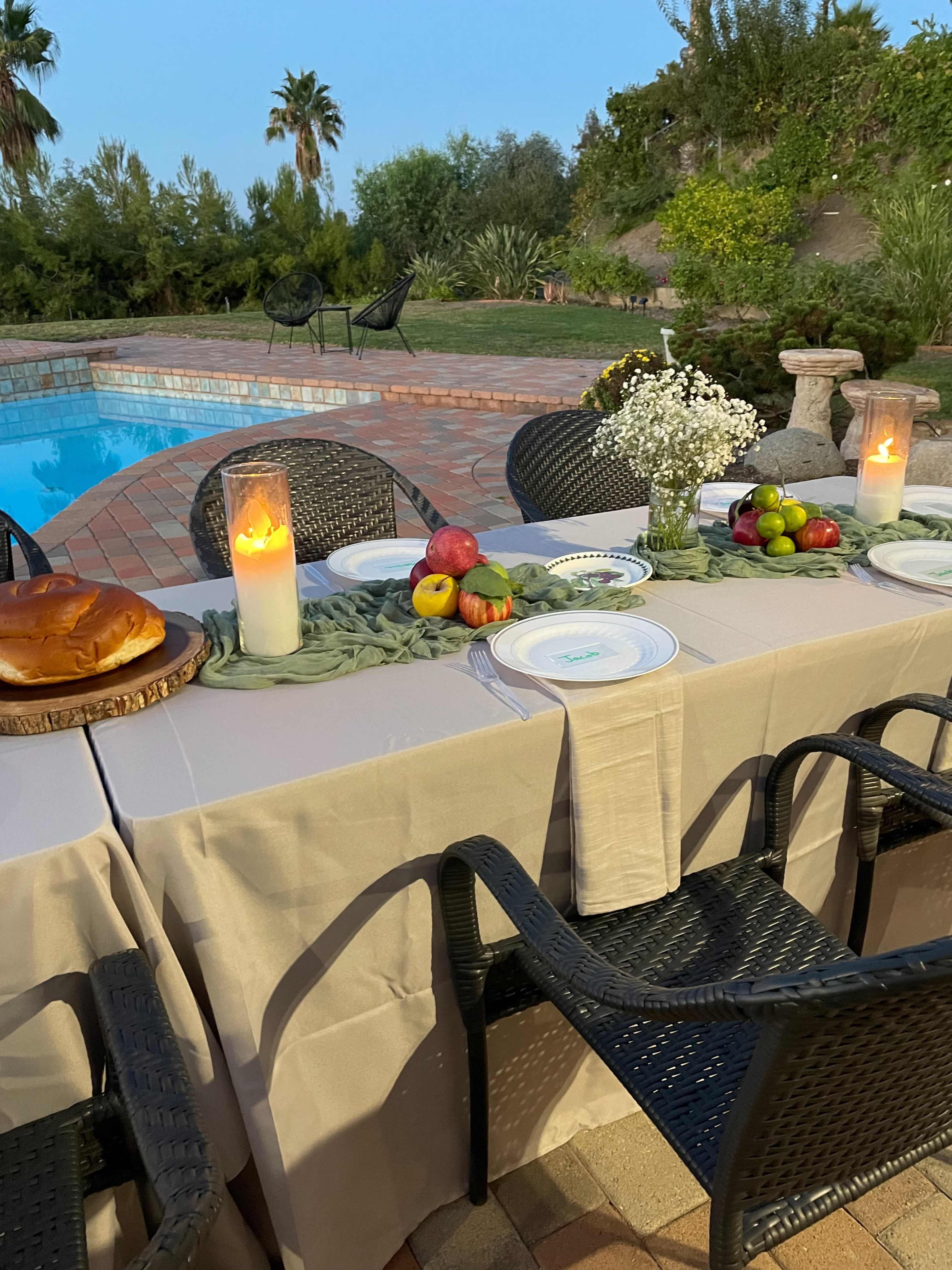 A table set with plates and fresh fruits is arranged beside a pool in a garden, with candles illuminating the scene at dusk.