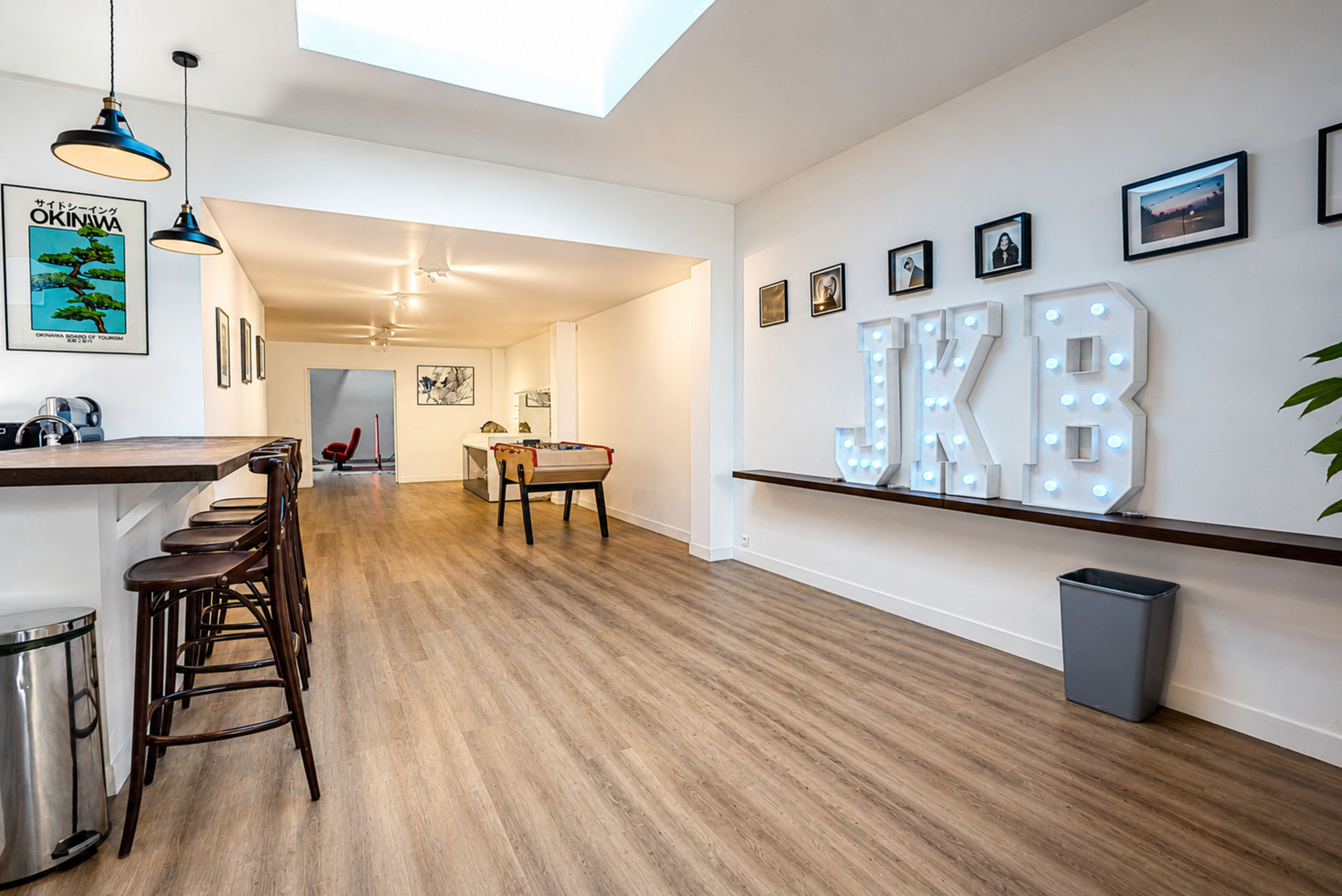 The image displays a modern, open-concept hallway featuring a bar area with stools, illuminated letters spelling "JKB," and framed photographs on the walls.