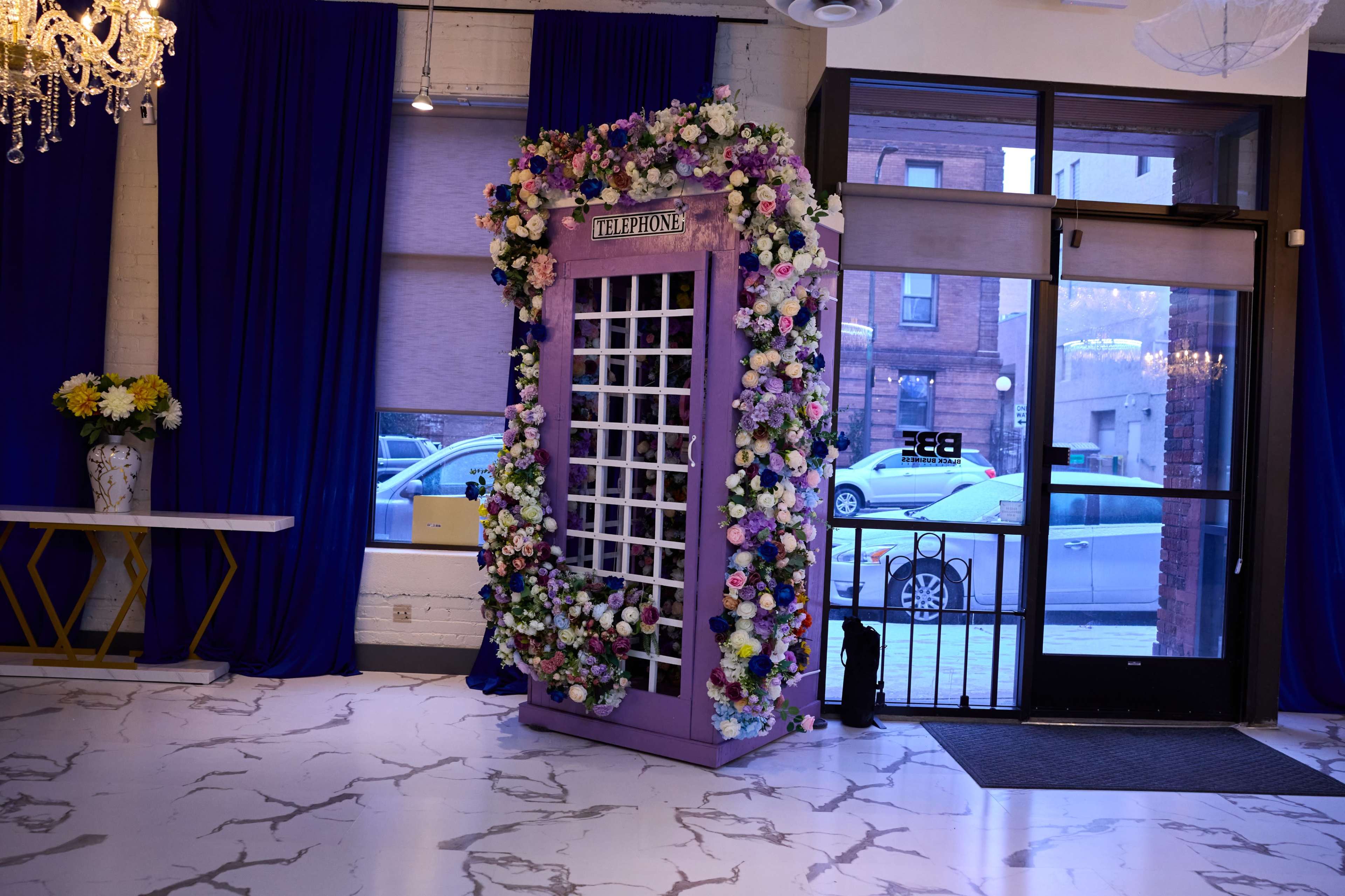 A large, floral-covered photo booth is positioned near a glass entrance in a decorated indoor space.