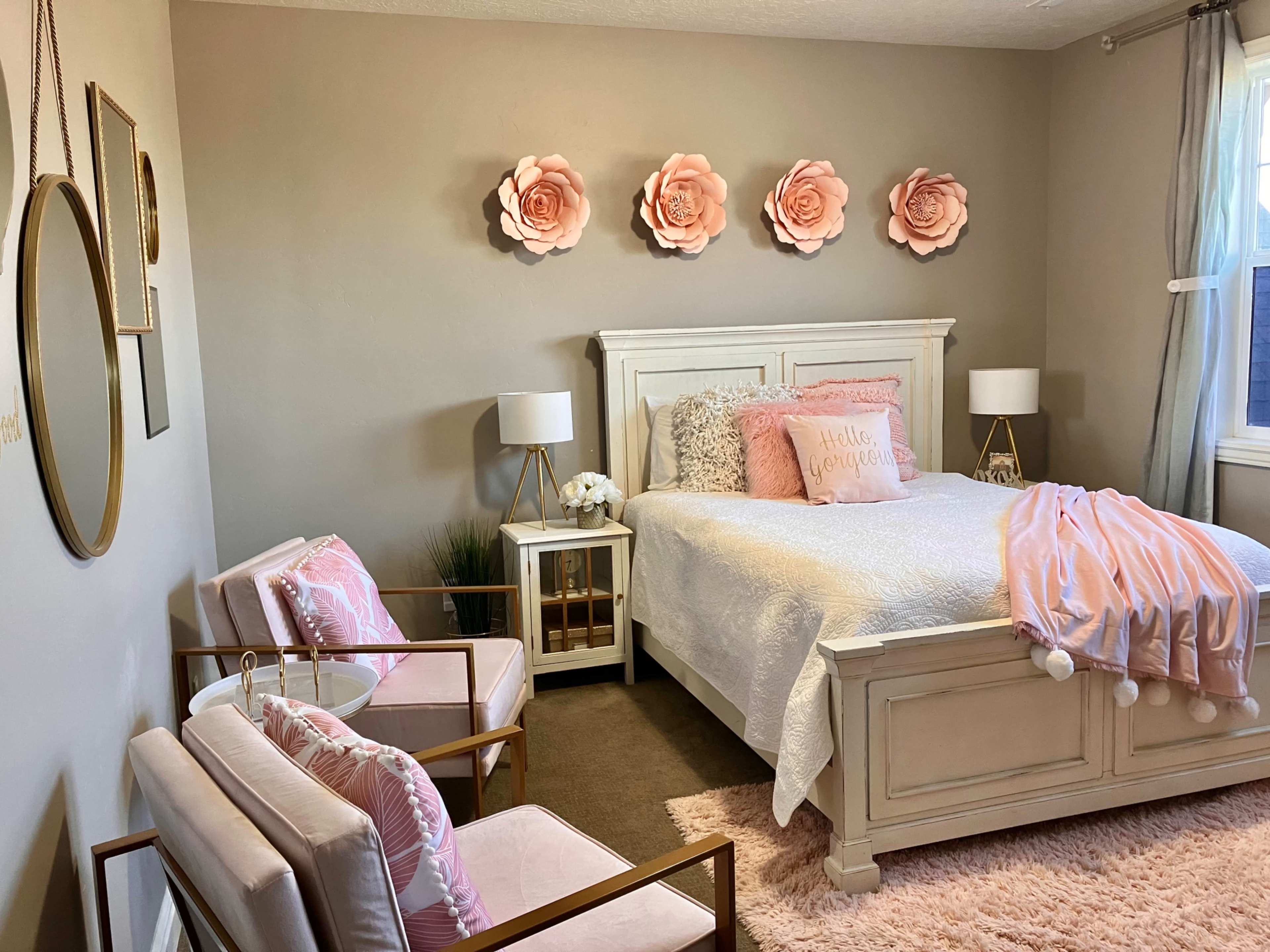 A well-decorated bedroom featuring a white bed with pink accents, two bedside lamps, a pair of armchairs, and large flower wall decorations.