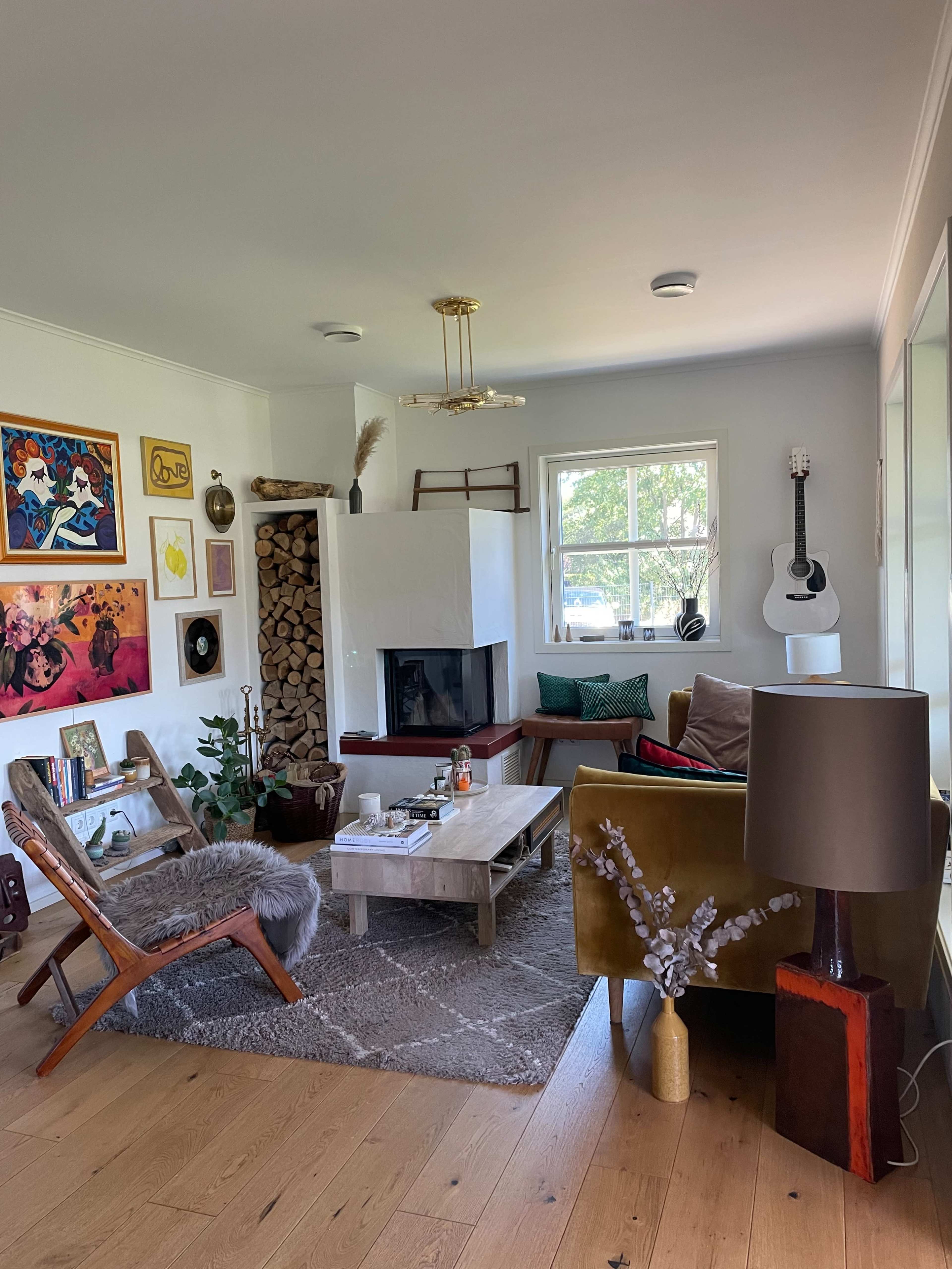 The image shows a cozy living room featuring a fireplace, a wooden coffee table, eclectic artwork on the walls, and a guitar in the corner.