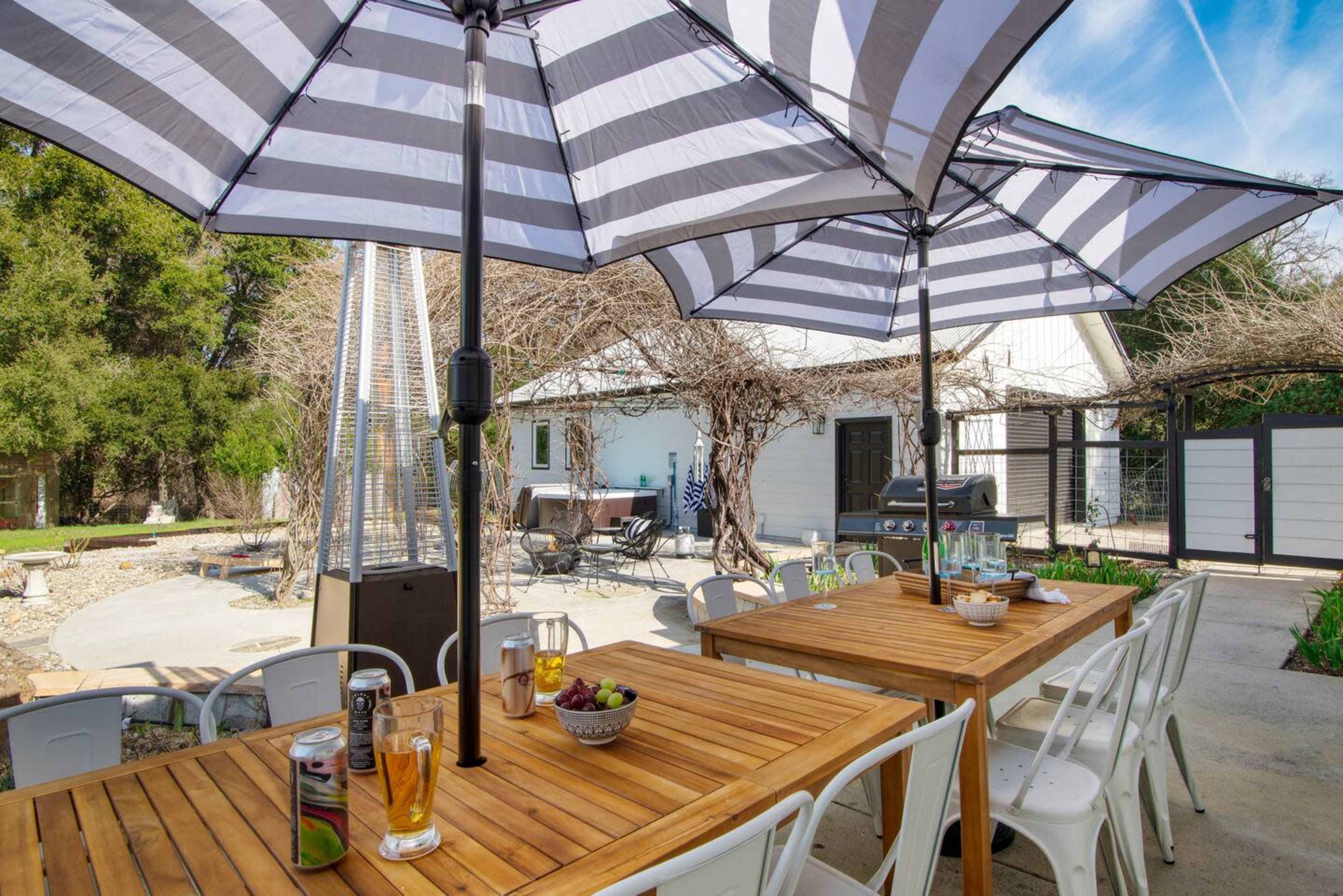 The scene features a spacious outdoor dining area with wooden tables, striped umbrellas, and a backdrop of a house surrounded by greenery.
