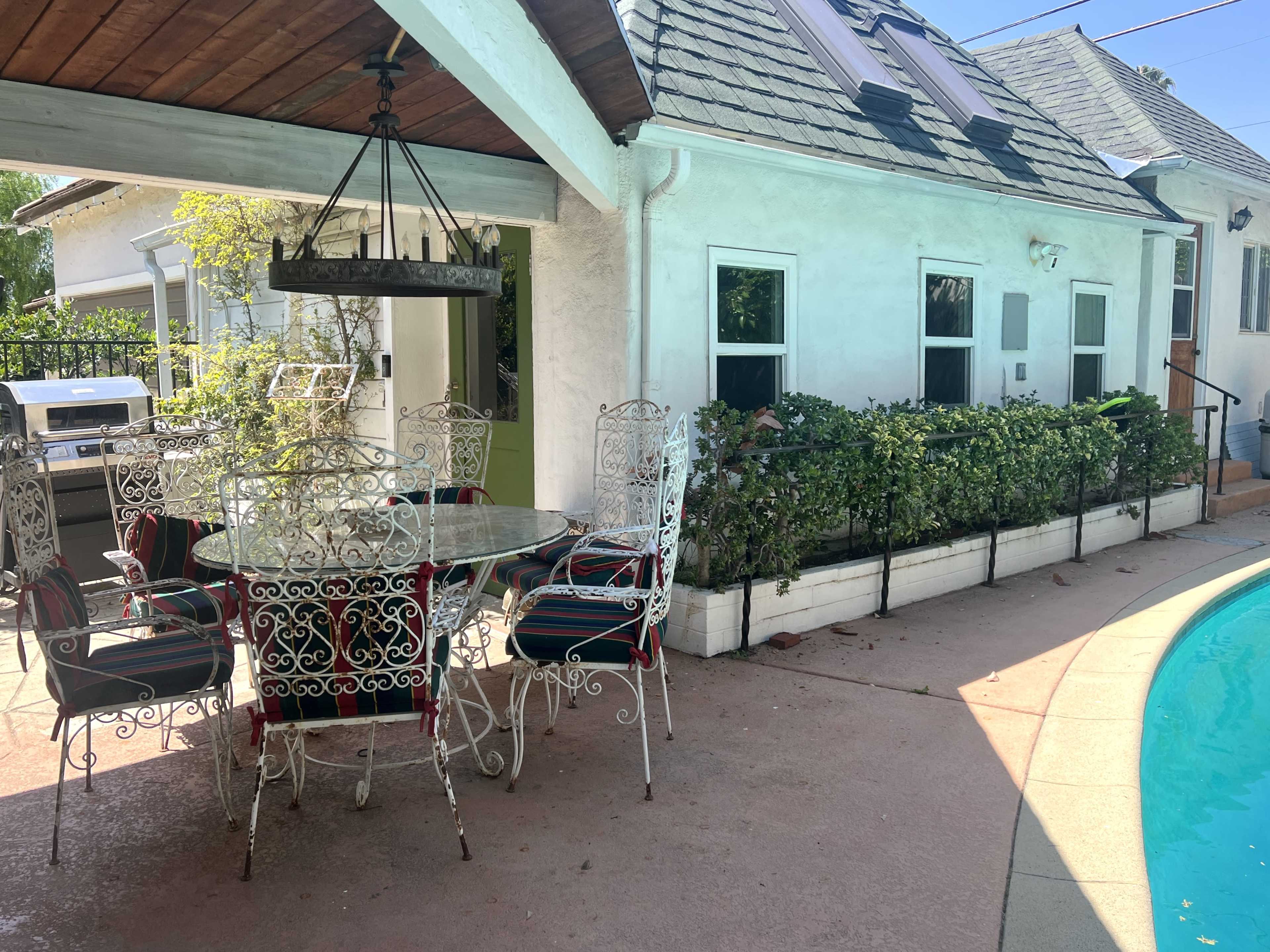 A circular glass table with ornate metal chairs is situated on a patio beside a swimming pool and a house with a sloped roof.