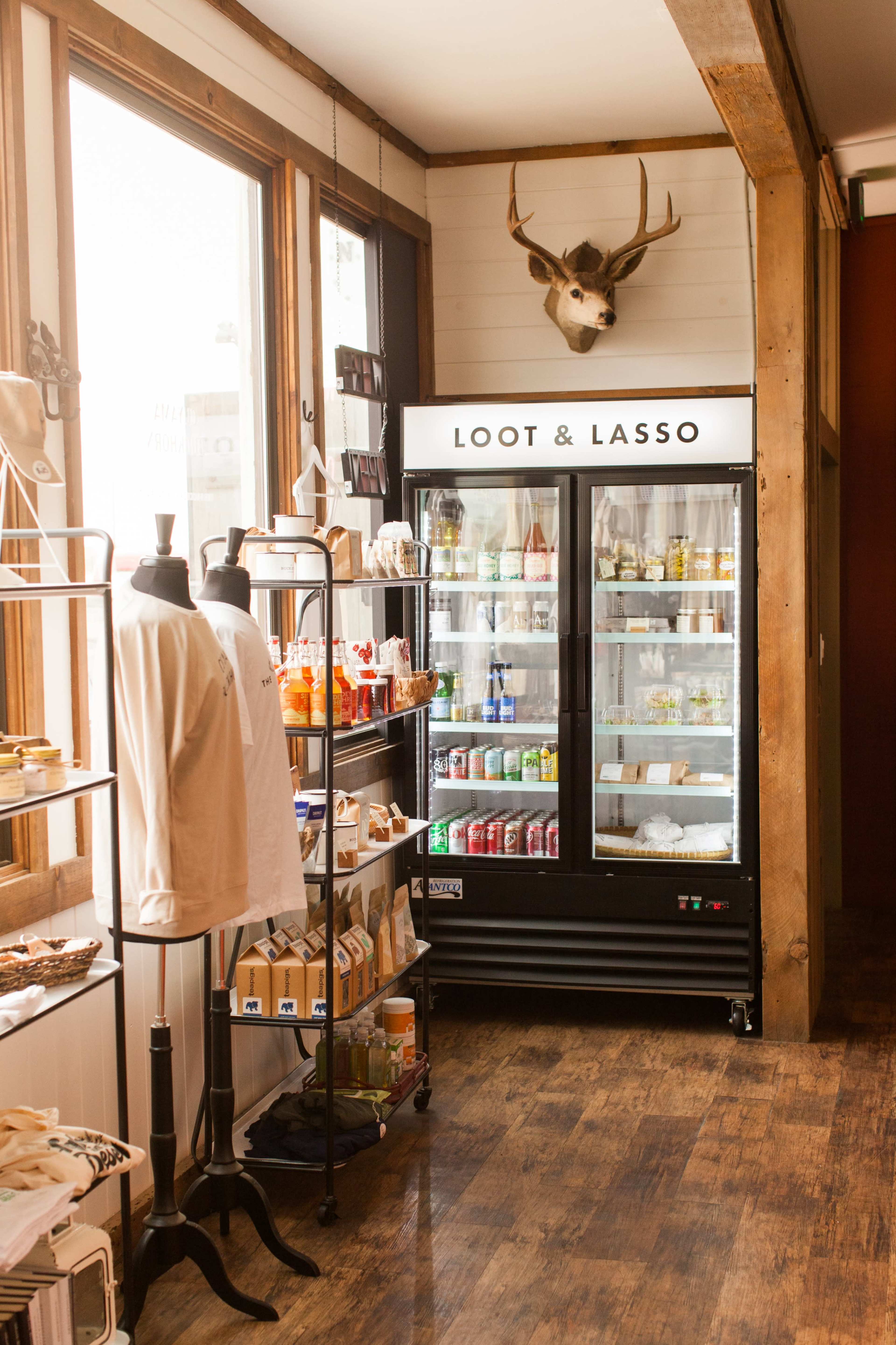 A corner of a shop featuring a refrigerator labeled "LOOT & LASSO," surrounded by shelves displaying various products and a mounted deer head above the refrigerator.
