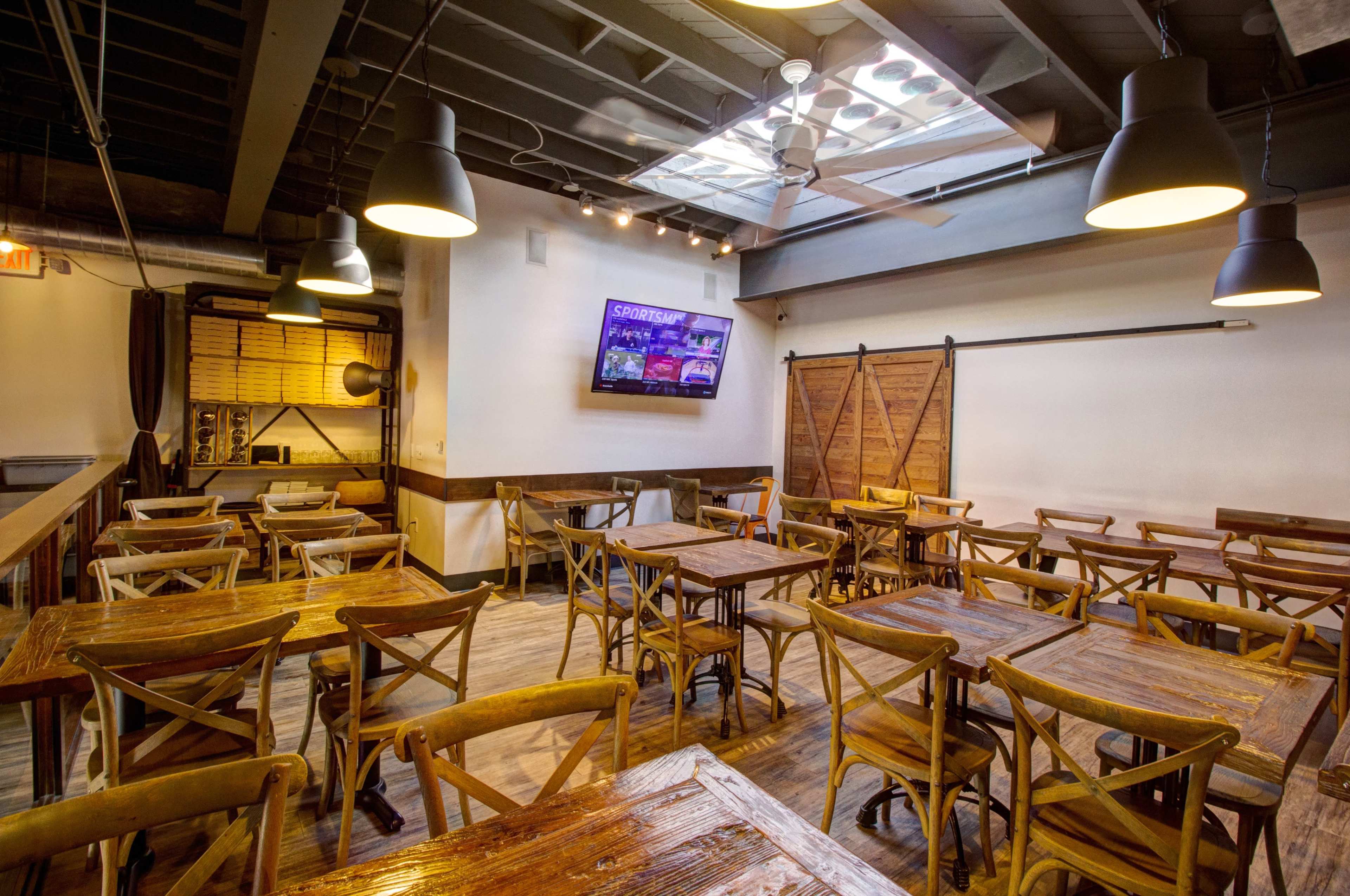 The image shows a rustic dining area with wooden tables and chairs, featuring a large television on the wall and a skylight overhead.