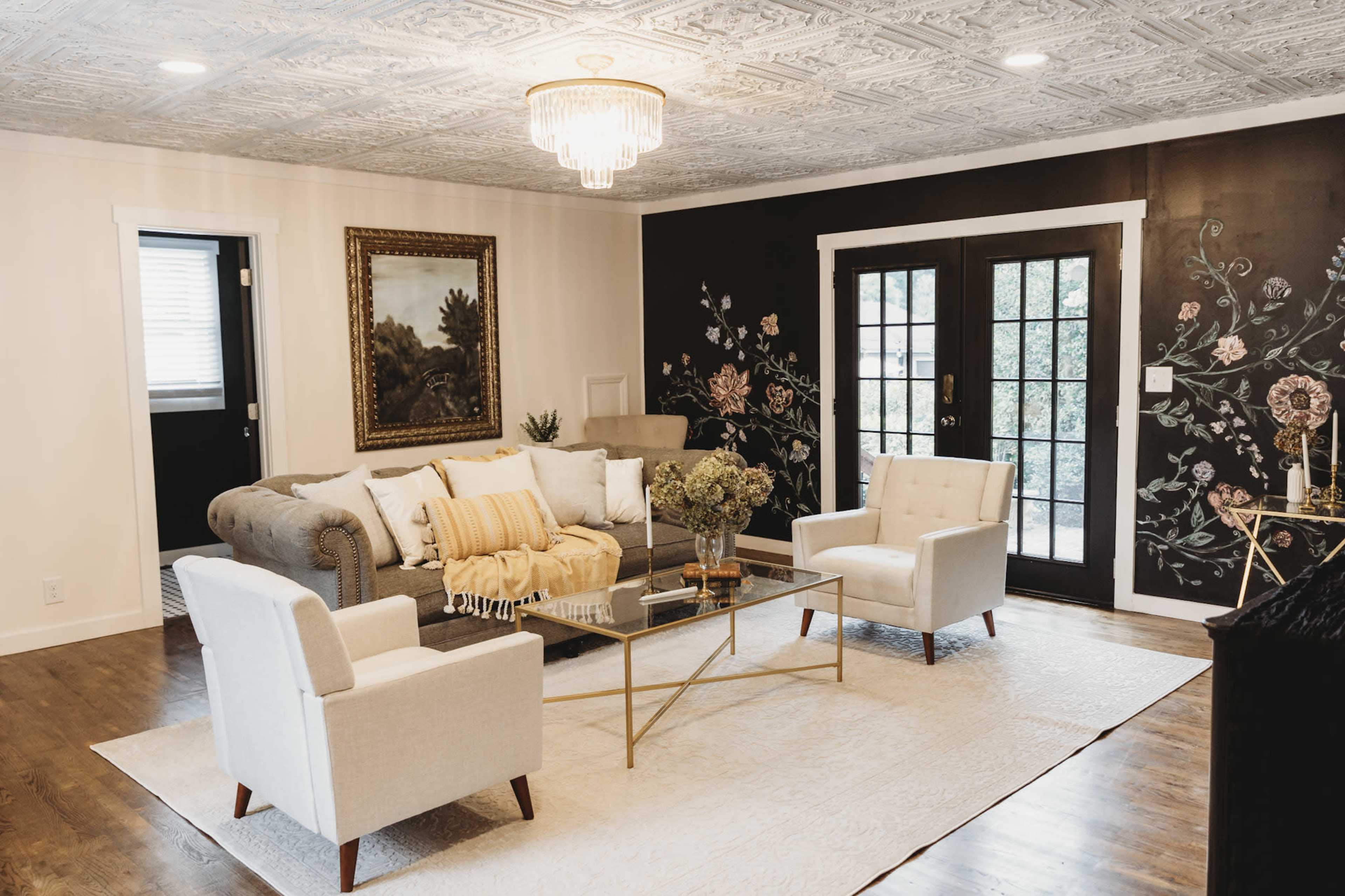 The image shows a living room featuring a gray sofa with decorative pillows, two white armchairs, a glass coffee table, and a floral wall mural.