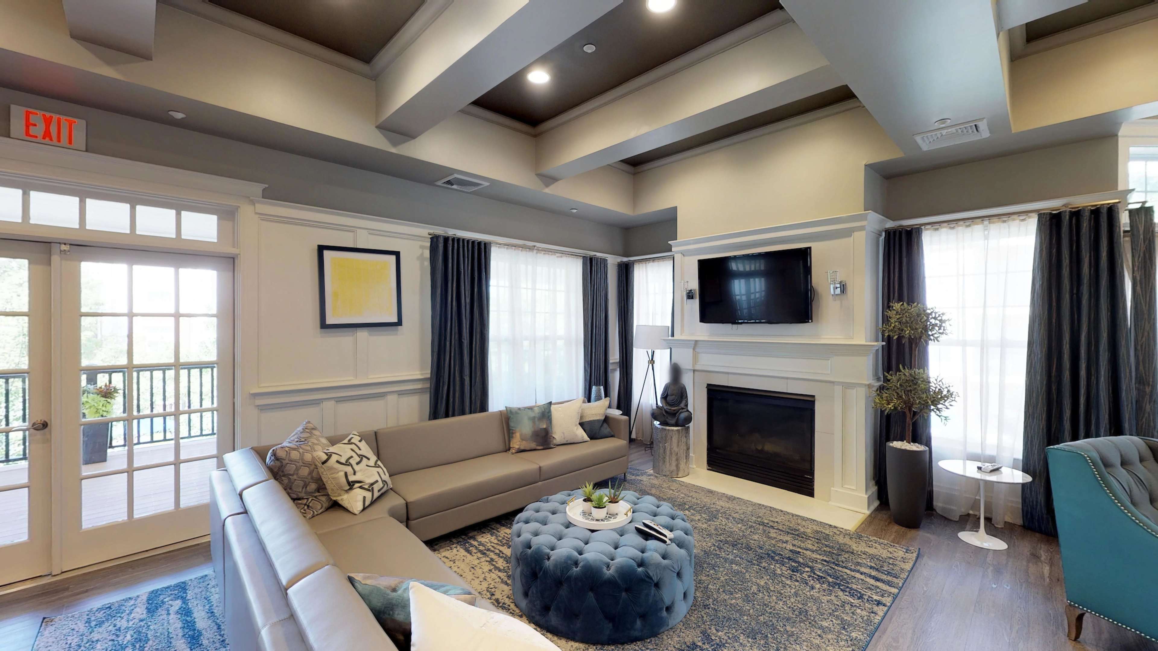 A modern living room with a gray sectional sofa, a blue tufted ottoman, a flat-screen TV mounted above a fireplace, and large windows with sheer curtains.