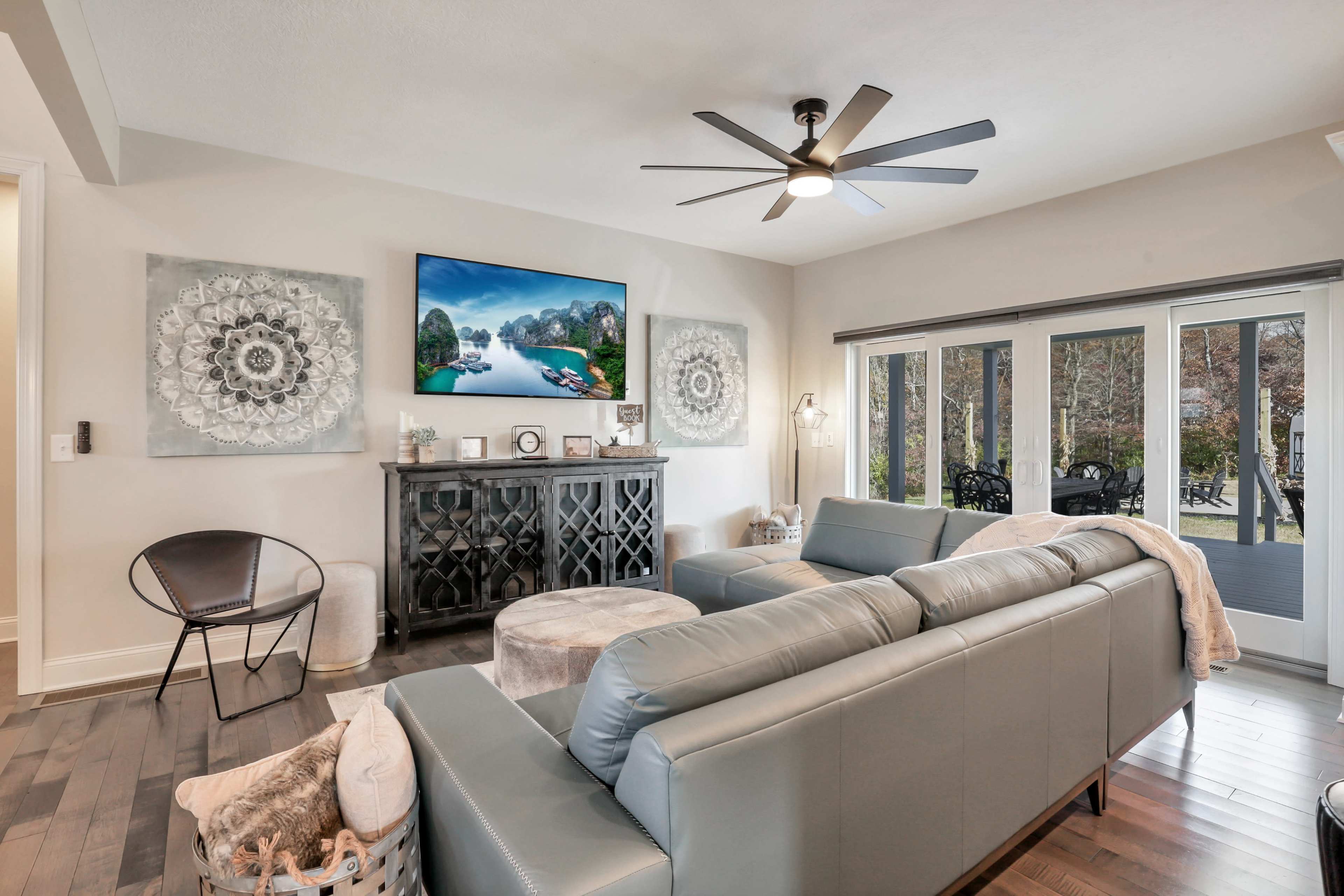 A modern living room features a sectional sofa, a circular ottoman, a sleek media console, and large windows that open to a patio.