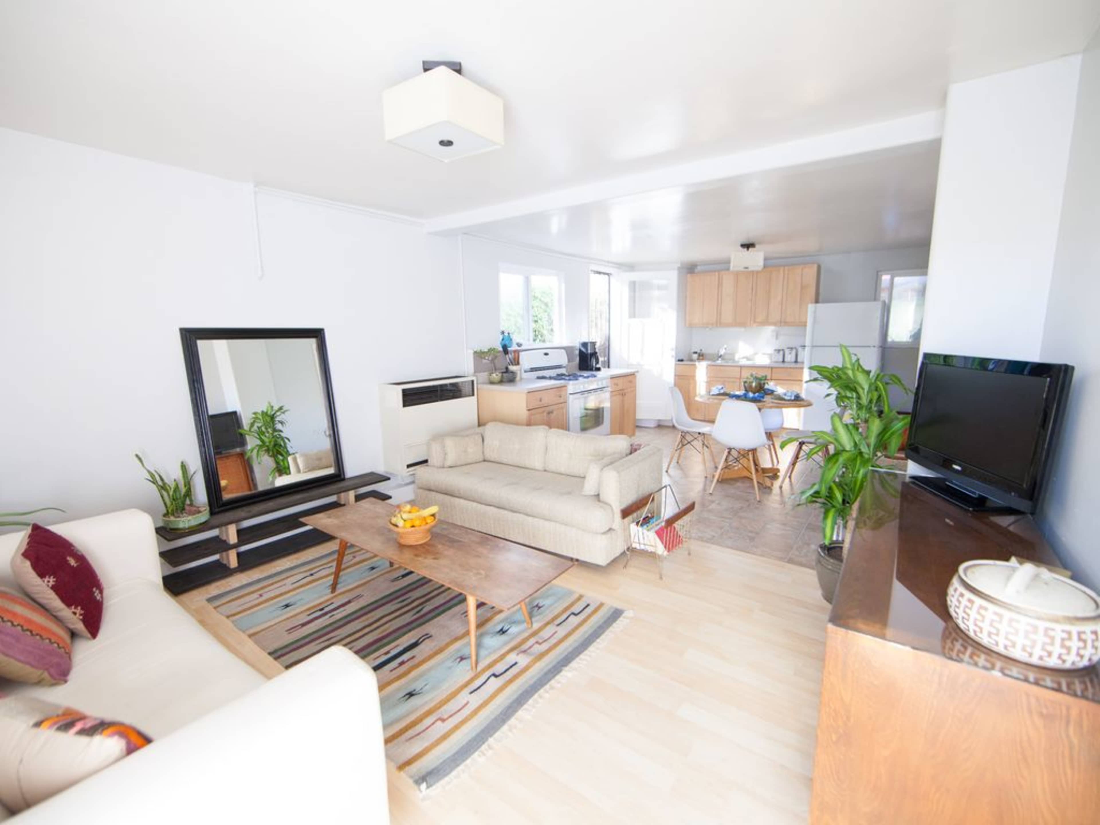 The image shows a bright living room with a sofa, a coffee table, a mirrored stand, and a kitchen area in the background.
