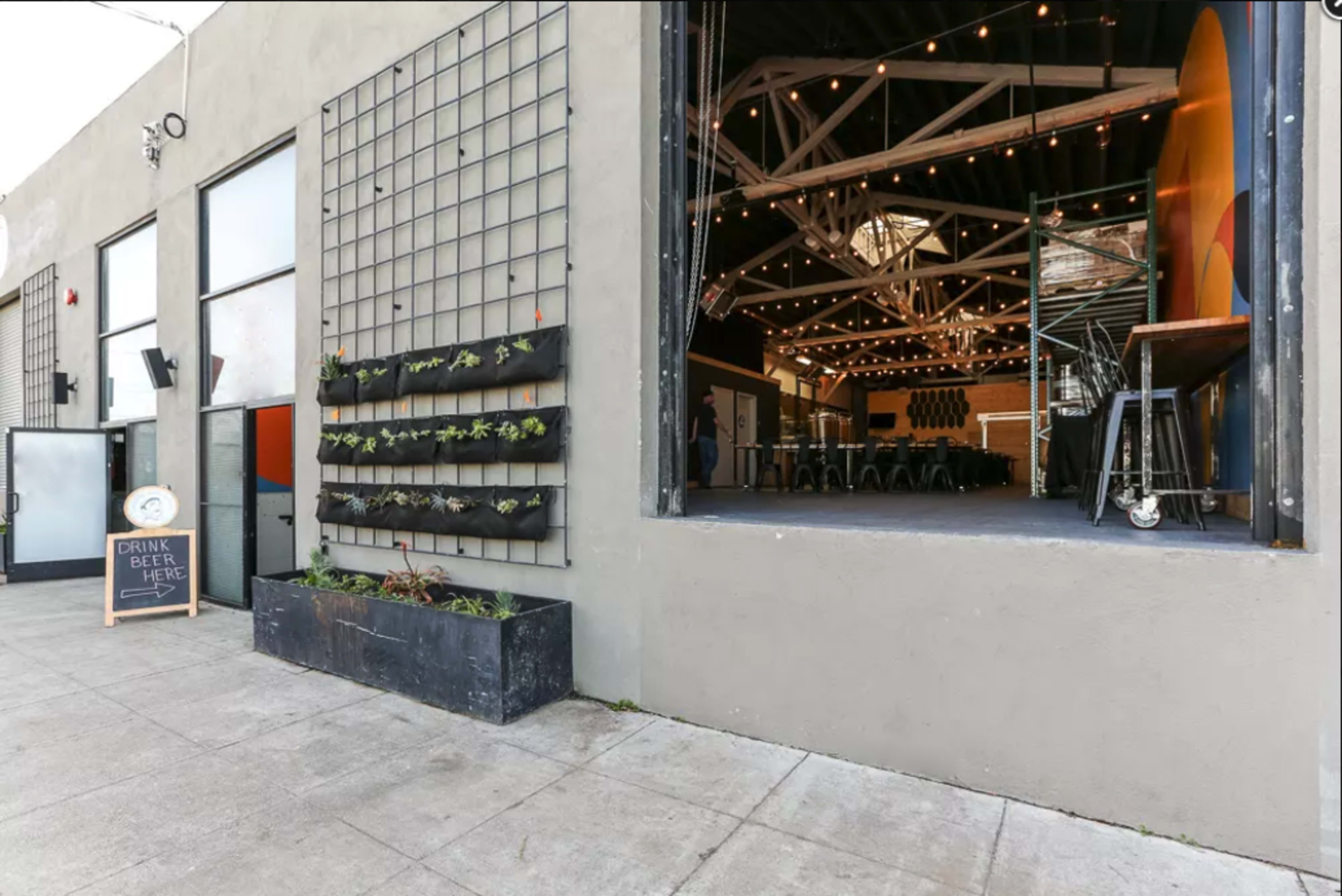 An industrial-style venue with a large open entrance, featuring a wall of plants and a sign that invites customers to enjoy drinks.