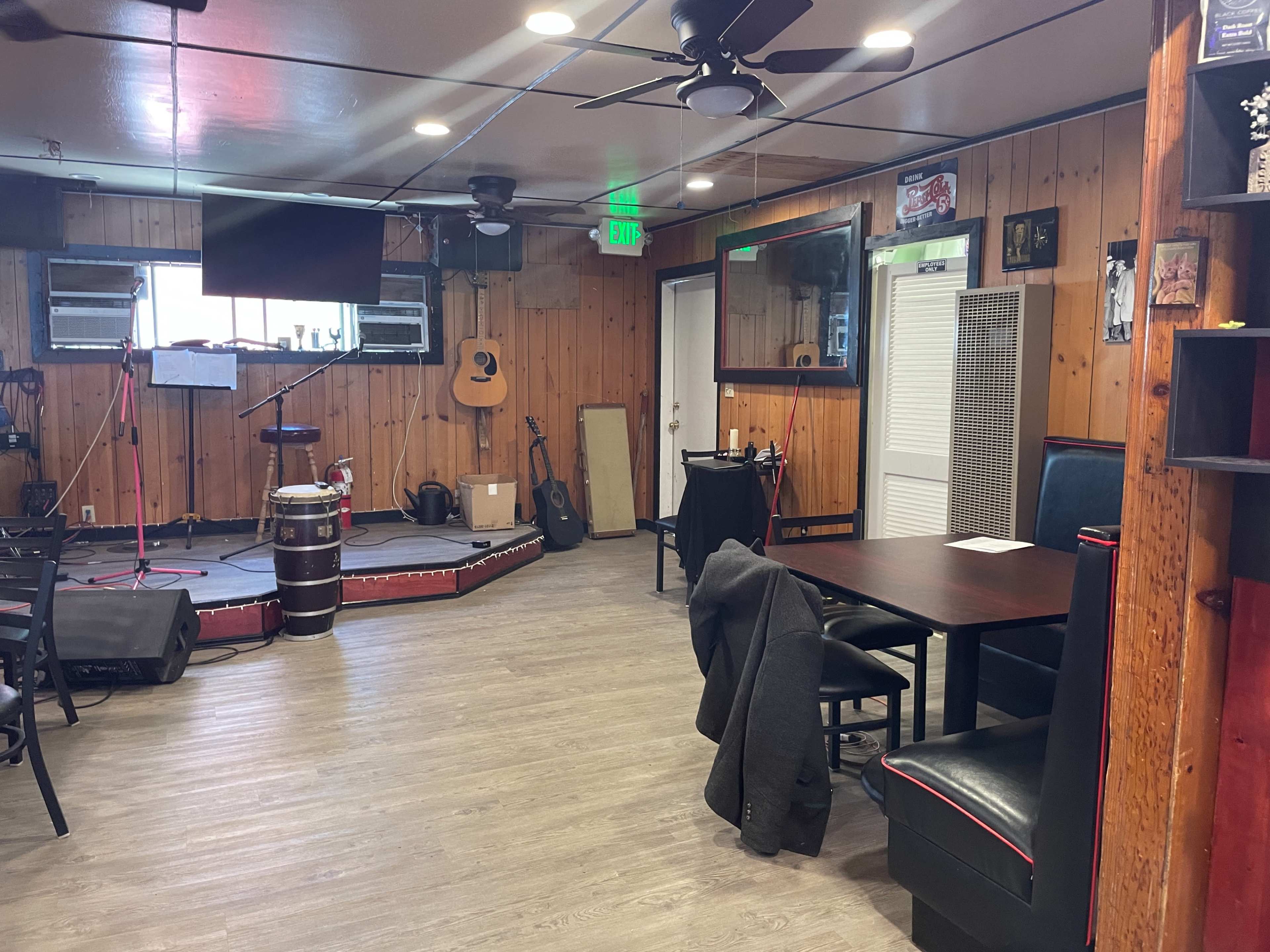 A rustic restaurant interior with wooden walls, a small stage for performances, a few tables, and a mix of chairs and benches.