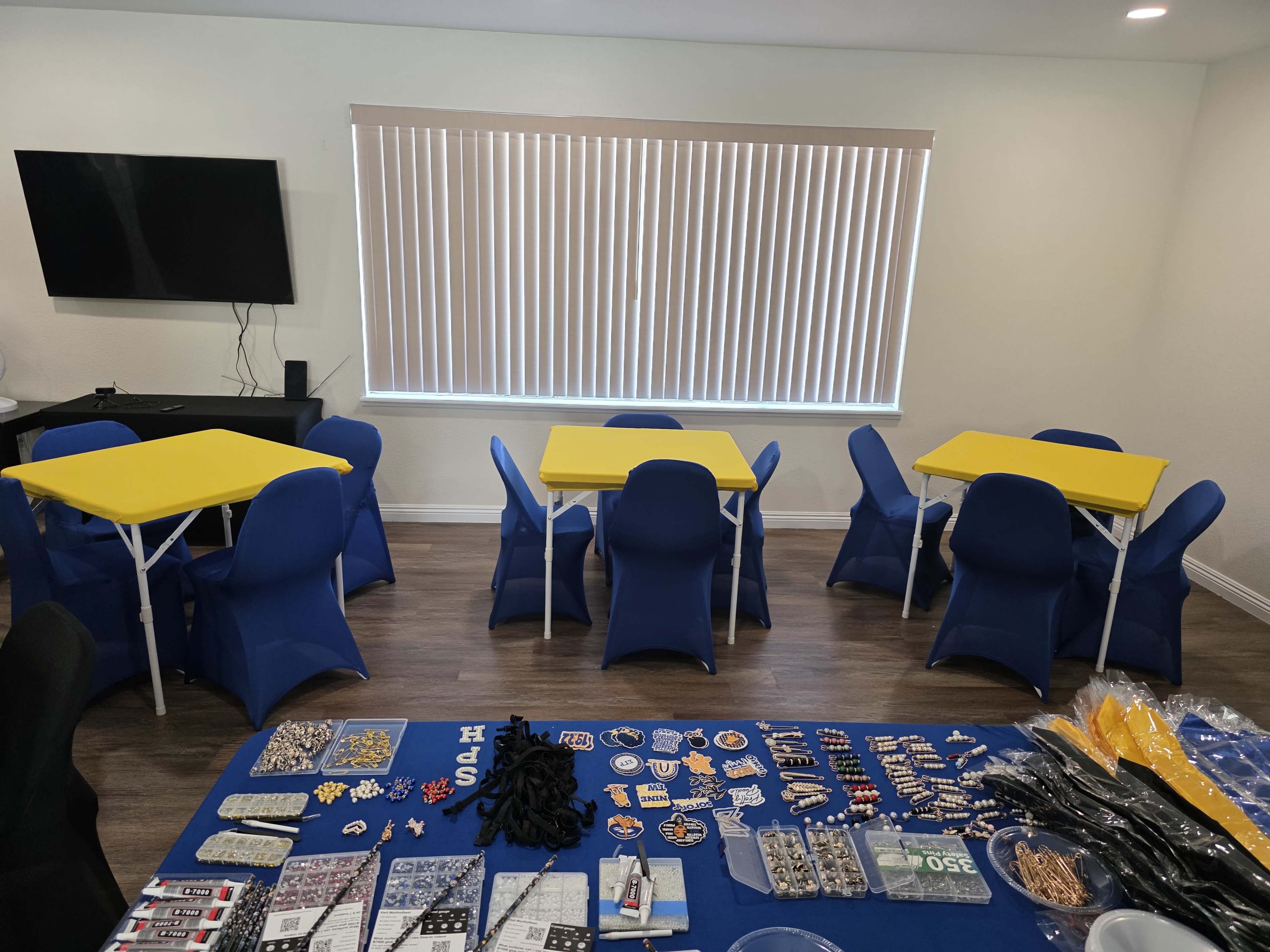 The image shows a brightly lit room with yellow tables and blue chairs arranged around the space, alongside a variety of items displayed on a blue tablecloth.
