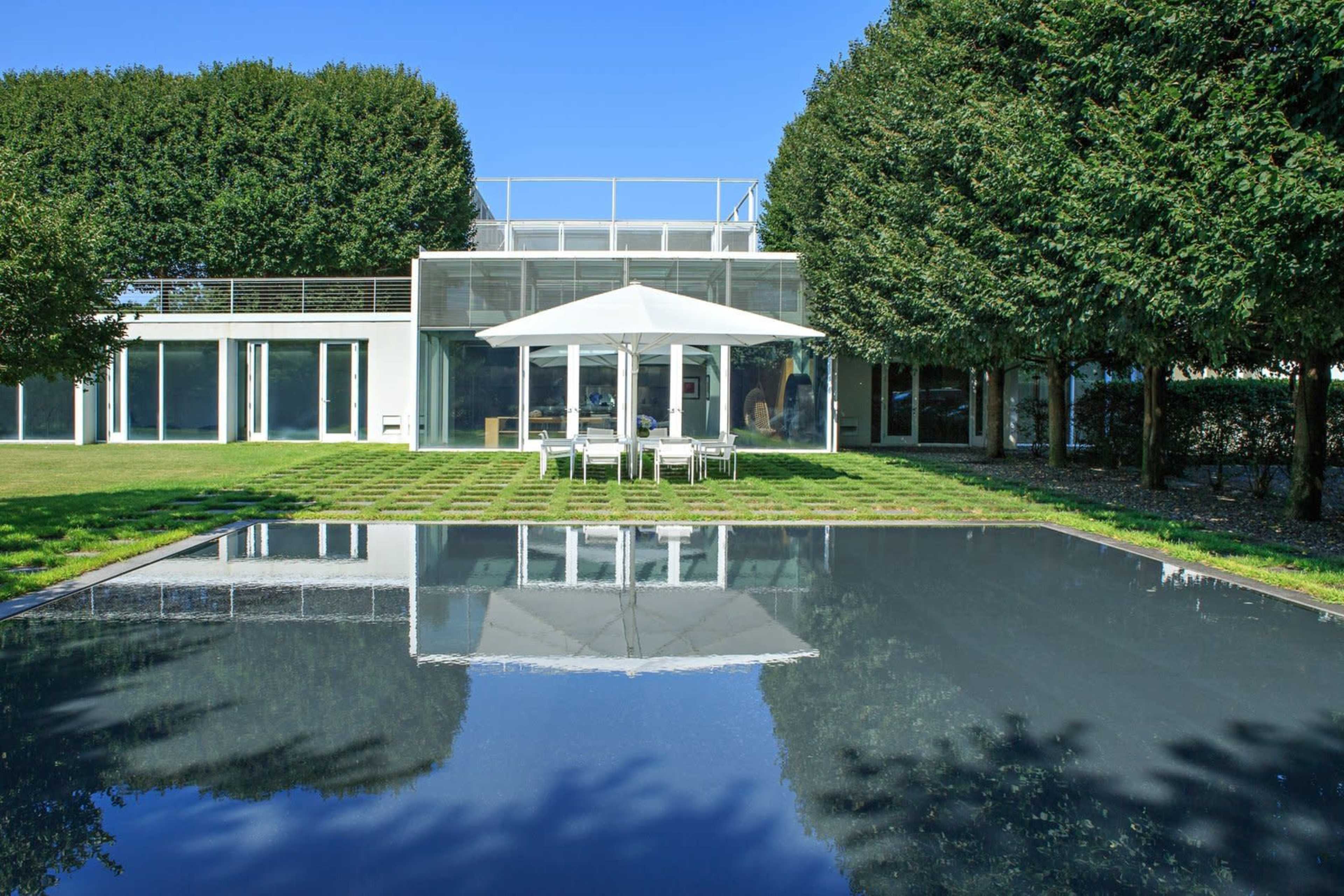 A modern house with large windows and a patio overlooks a rectangular swimming pool surrounded by neatly trimmed hedges.