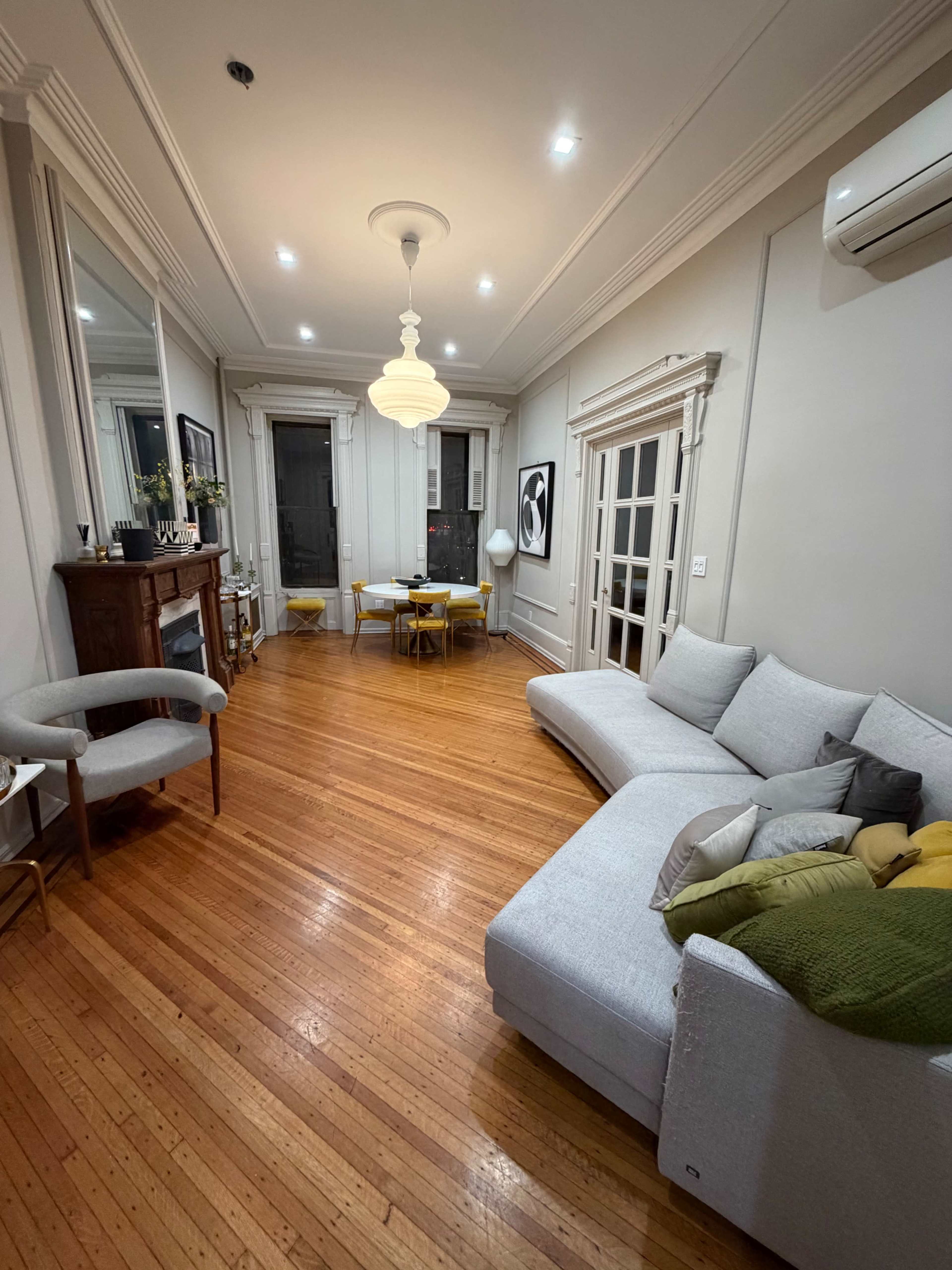 The image shows a spacious living room with a light-colored sectional sofa, wooden flooring, and a dining table set for four.