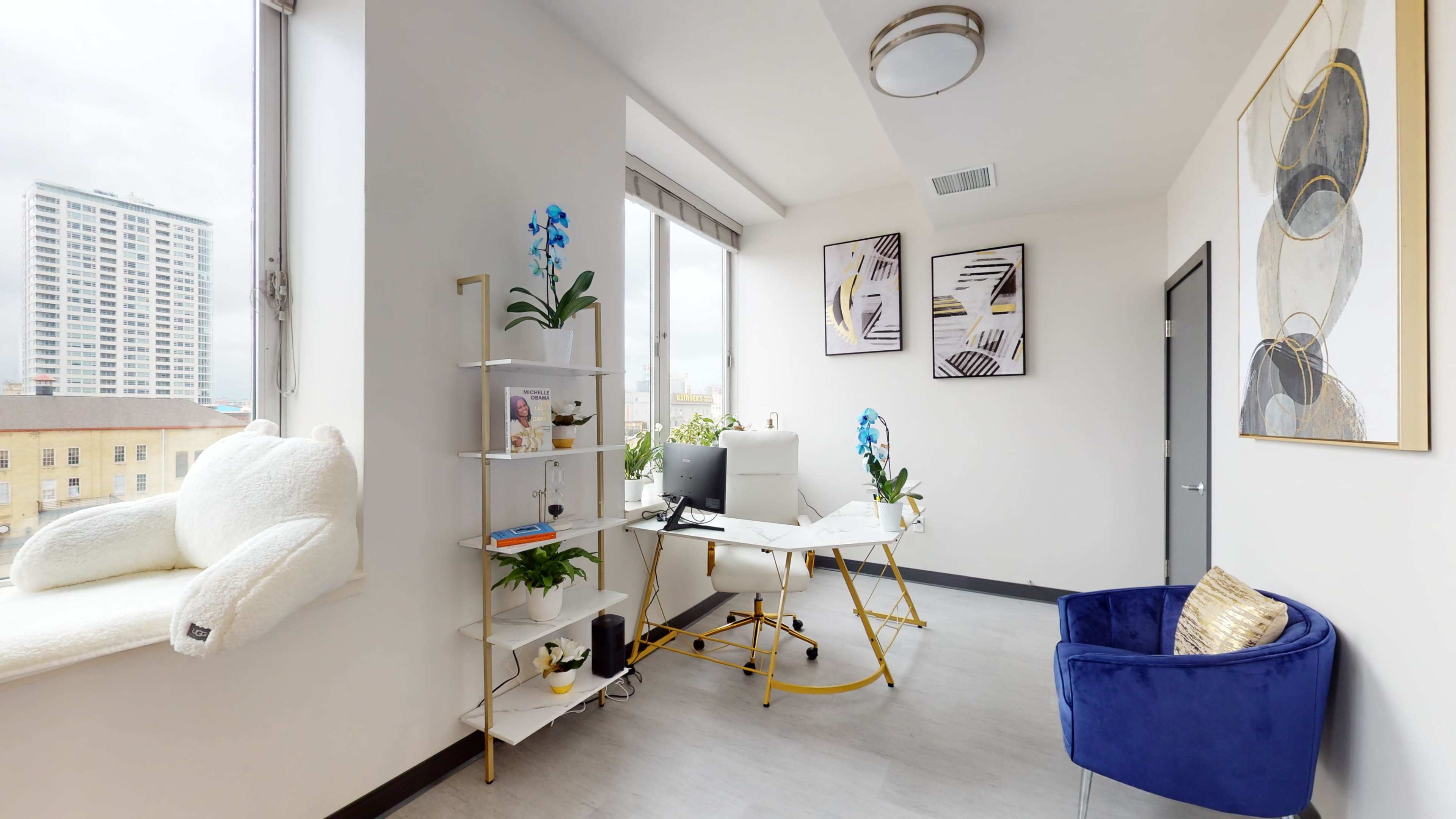 A bright office space features a desk with a computer, plants, and wall art, along with a cozy seating area by a large window.