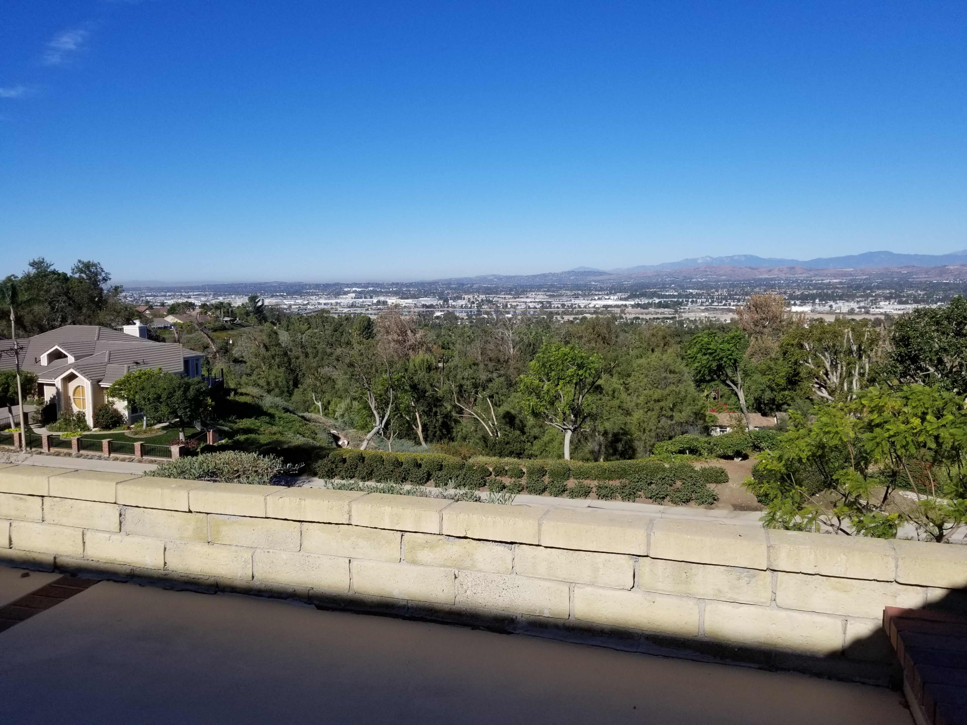 The image shows a view overlooking a cityscape surrounded by trees and hills under a clear blue sky.