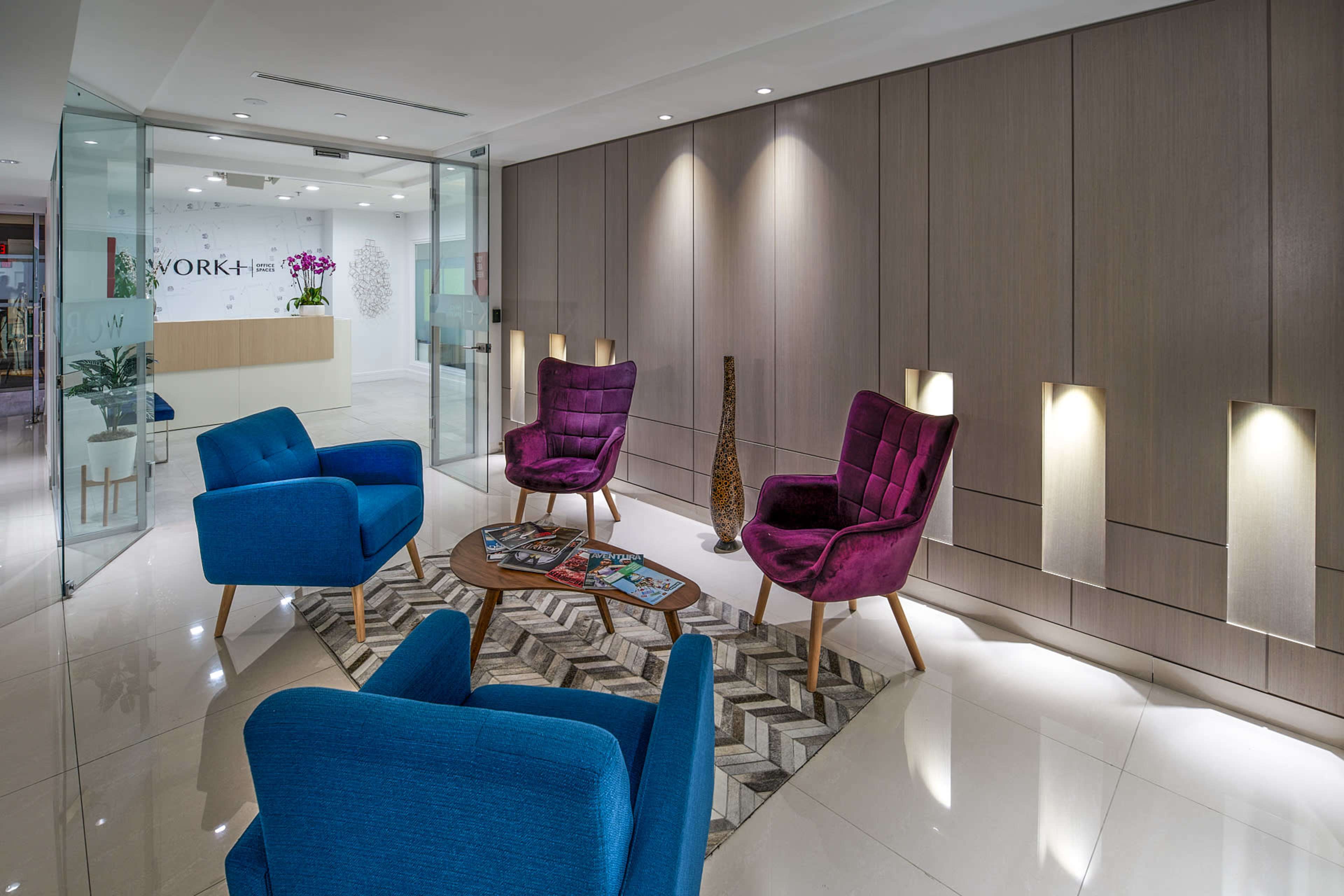 A modern reception area featuring blue and purple armchairs, a round table with magazines, and a glass partition leading to a front desk.