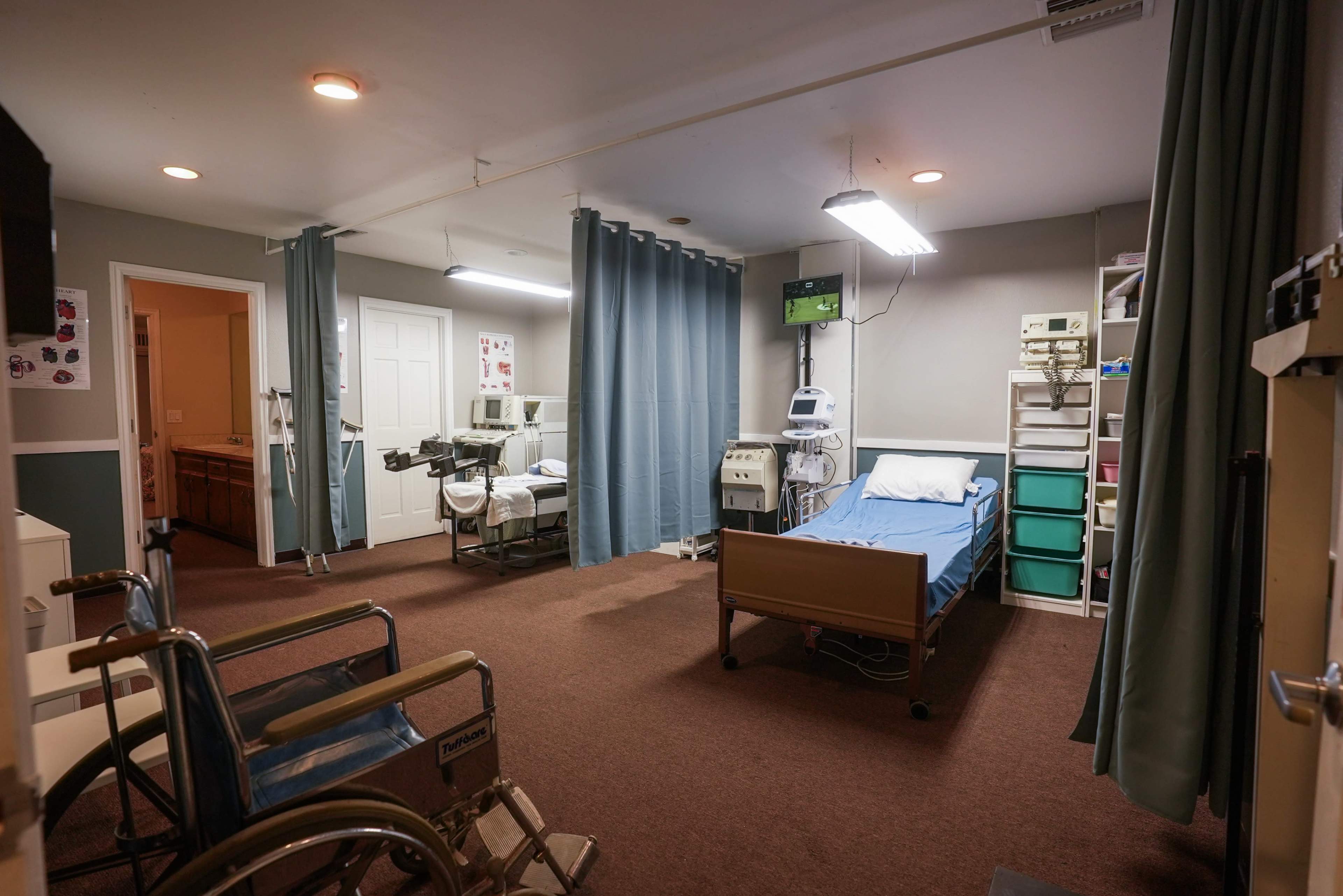 The image shows a hospital room with two beds, medical equipment, and wheelchairs arranged along the walls, separated by curtains.
