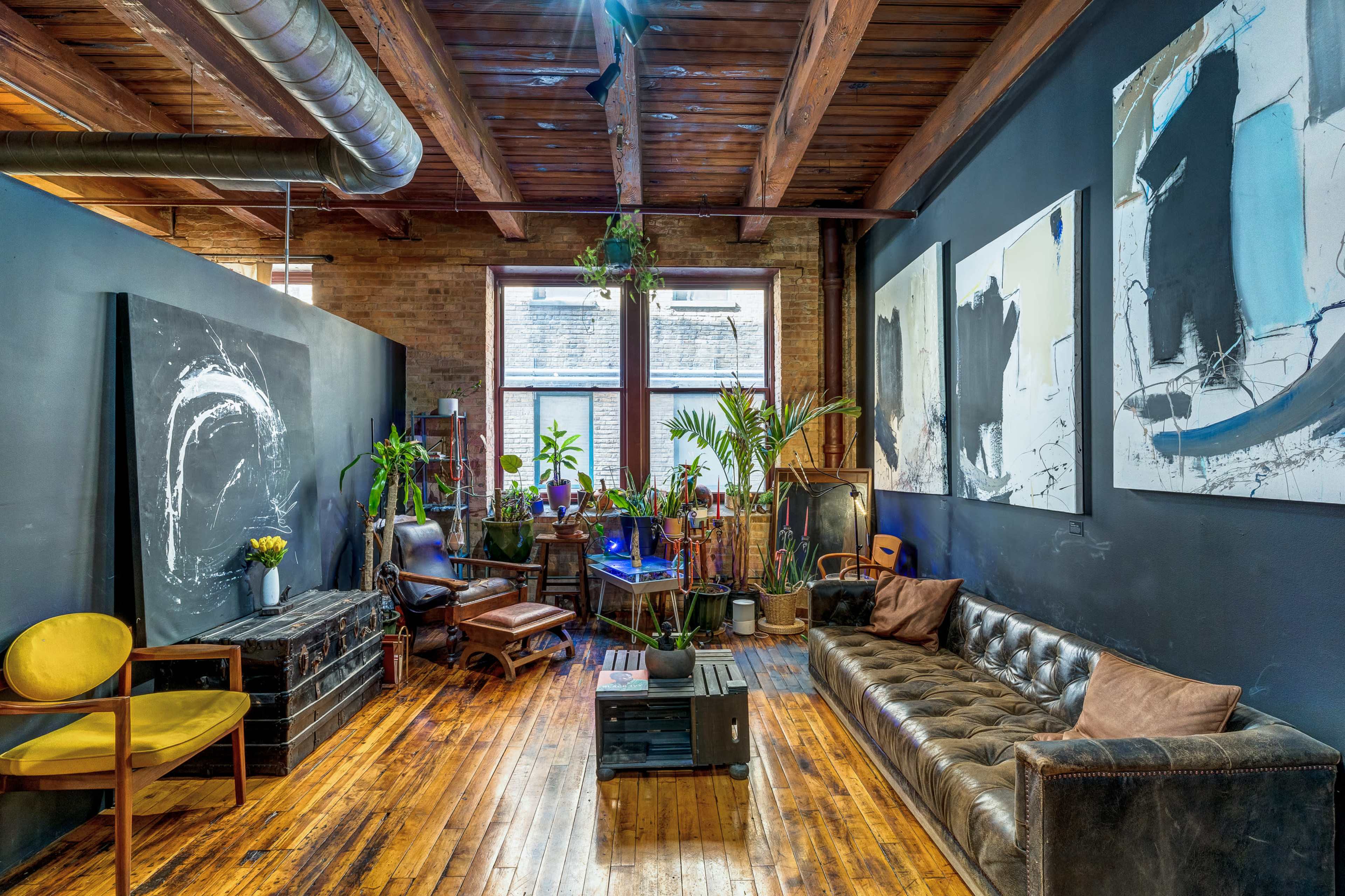 A cozy room with wooden floors, a leather sofa, various plants, and artwork on the walls, all under exposed wooden beams.
