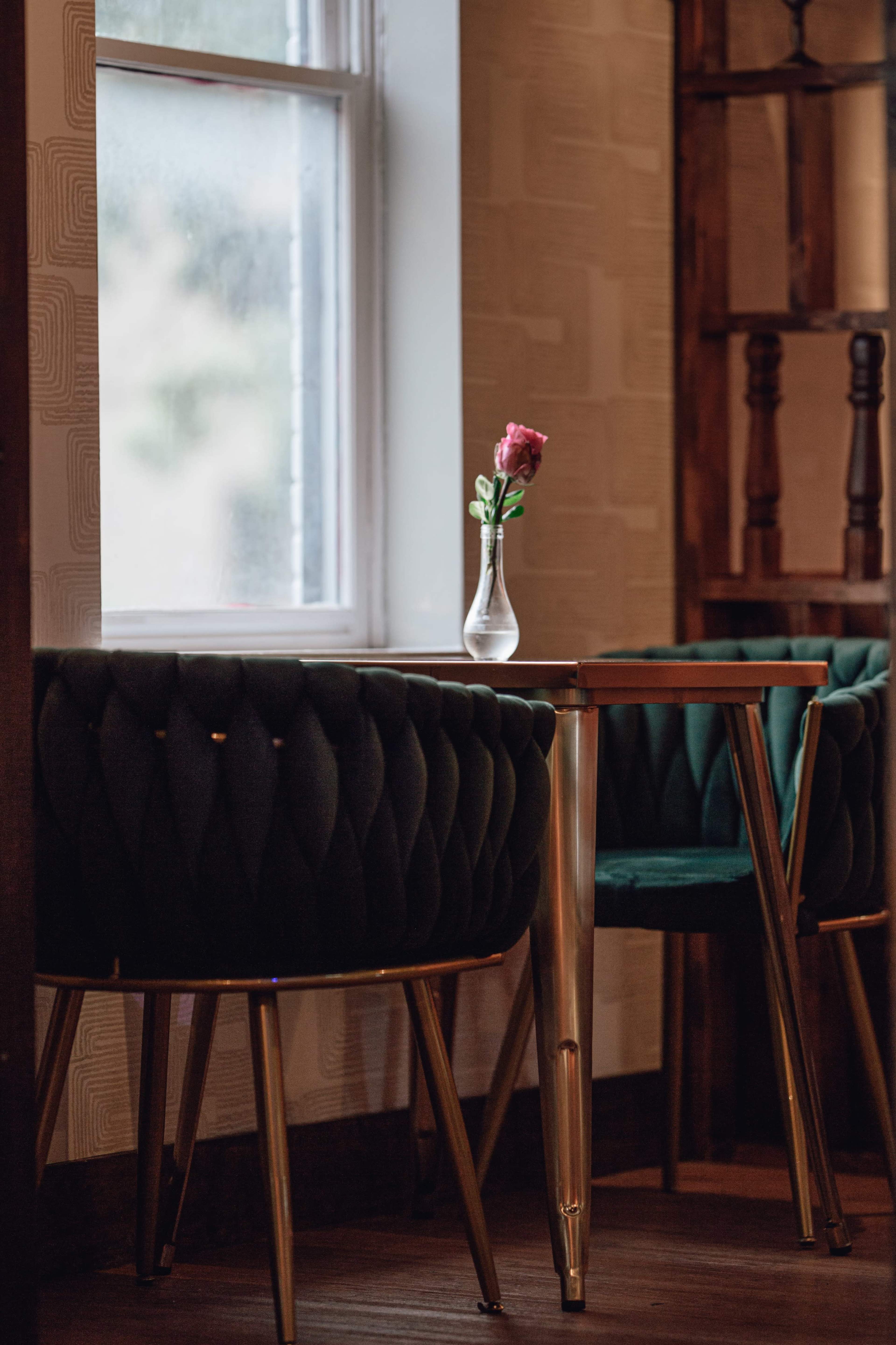 A small table with a glass vase containing a single rose stands beside a green, tufted chair in a softly lit room.
