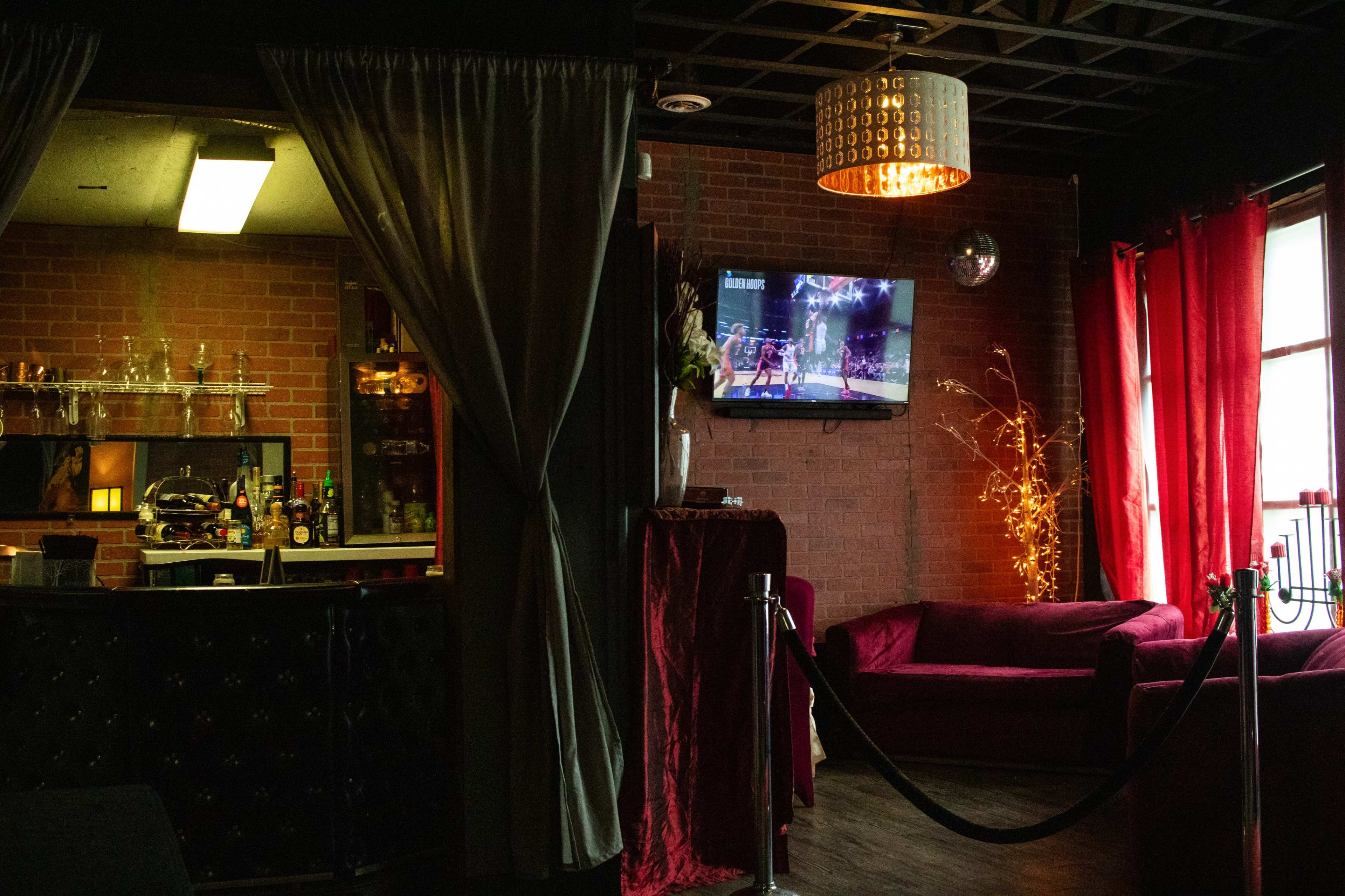 A dimly lit lounge area with a bar, a television displaying a sports event, and plush red seating surrounded by curtains.