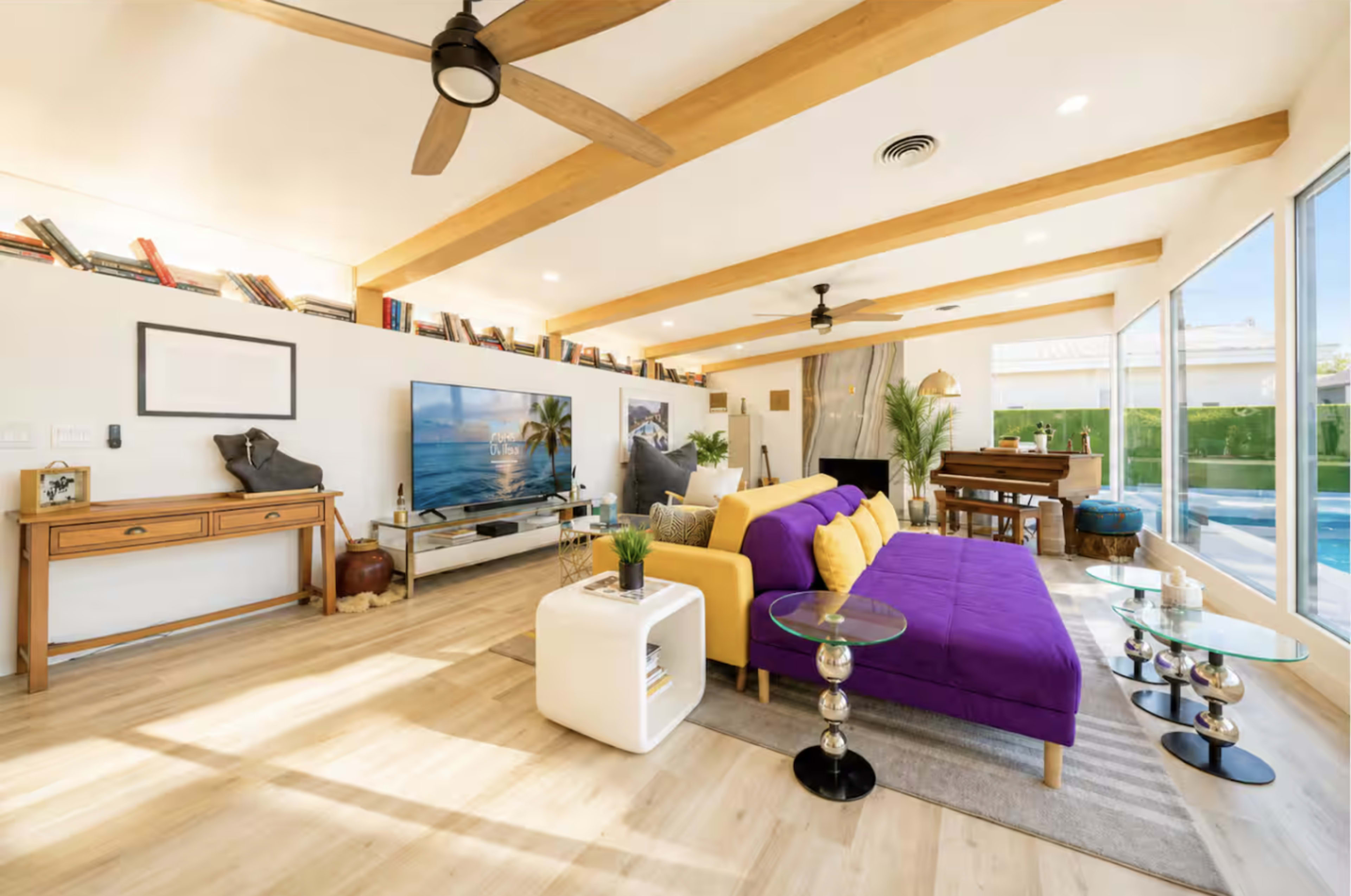 The living room features a yellow and purple sofa, a television mounted on the wall, and large windows overlooking a pool area.