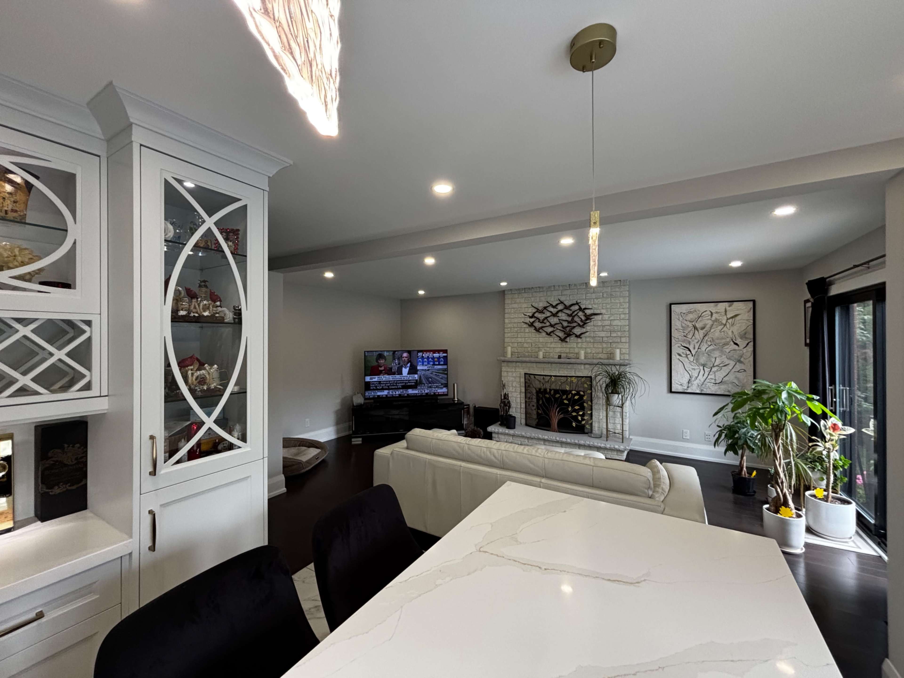 The image shows a modern living room featuring a white sofa, a fireplace with an ornate design, a television mounted on the wall, and a dining area with a marble countertop.
