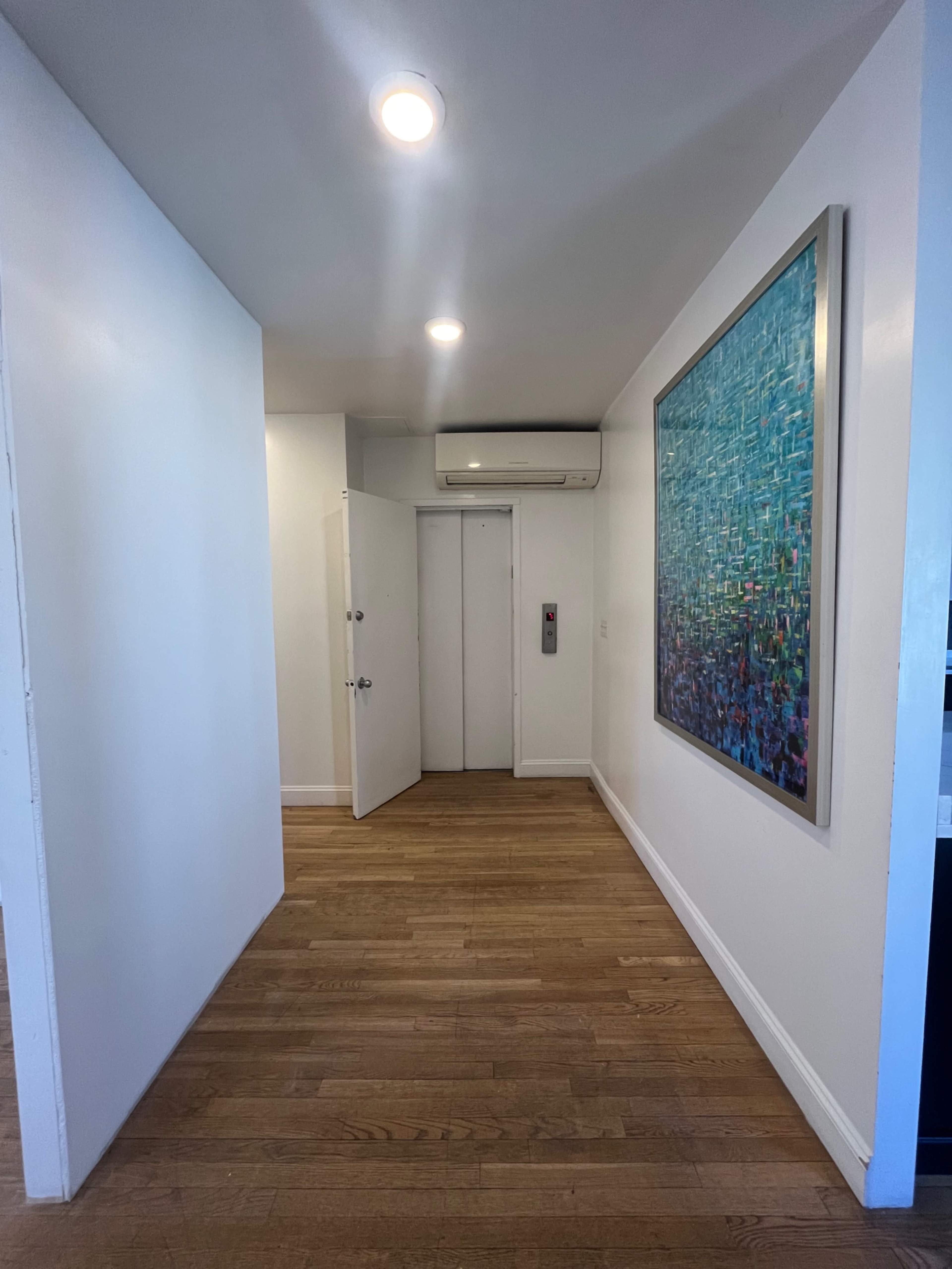 The image shows a hallway with wooden flooring, leading to a set of closed doors and a large piece of abstract art on the wall.