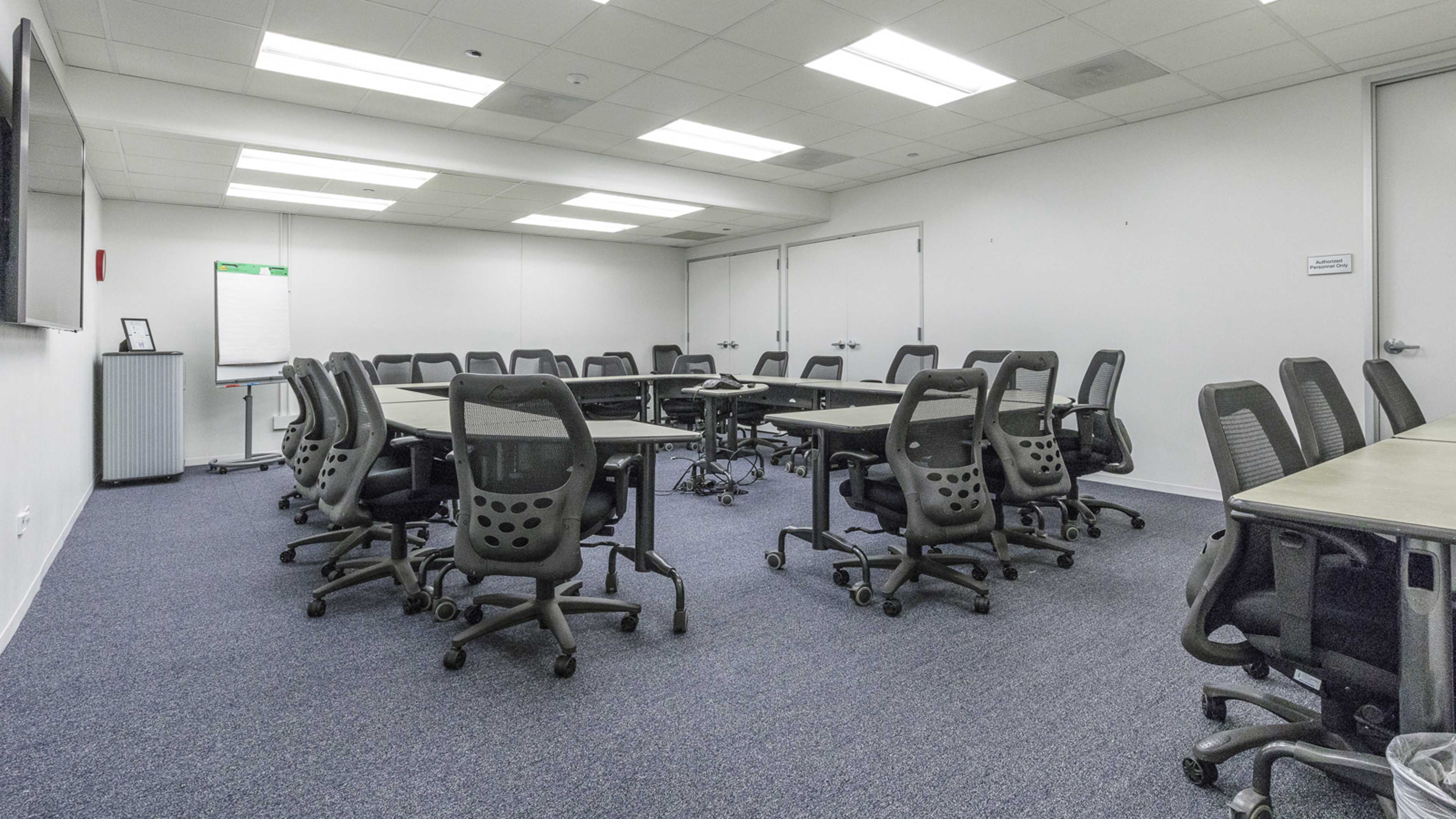 A large conference room is arranged with multiple tables and chairs, featuring a whiteboard and a television at one end.