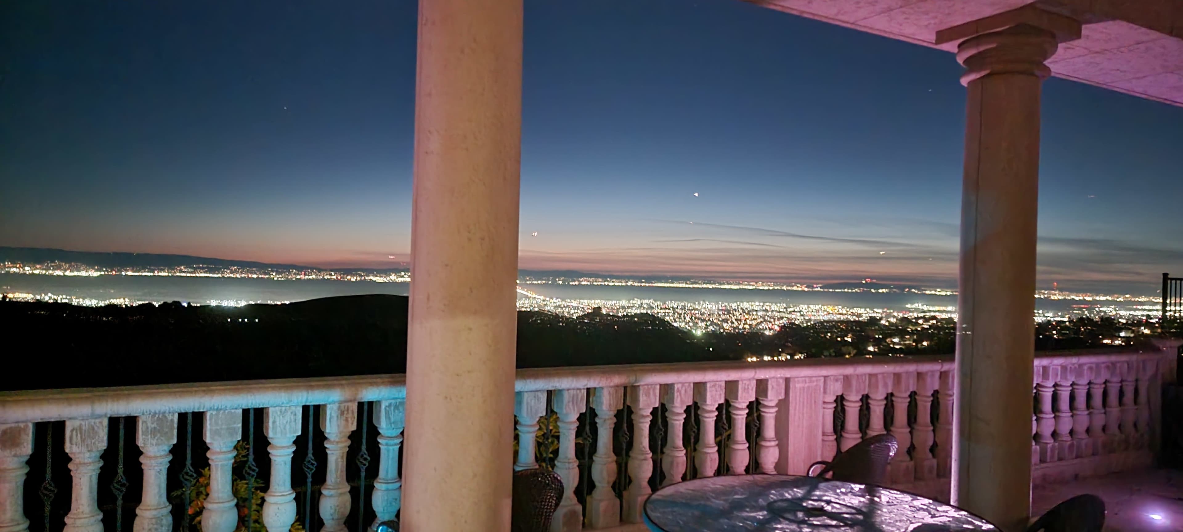 The image shows a balcony with a stone railing overlooking a city illuminated by lights, under a dusk sky transitioning from blue to orange.