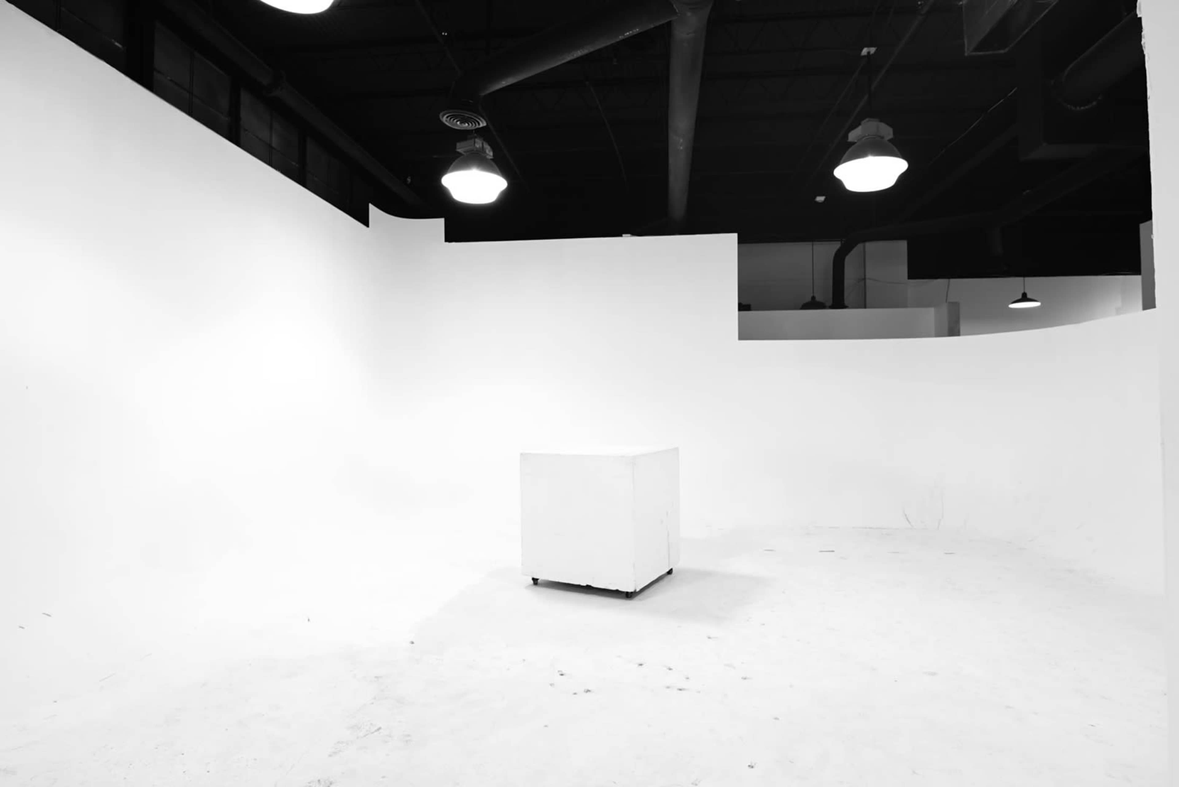 A vacant, white room with a single white cube in the center on a light-colored floor.