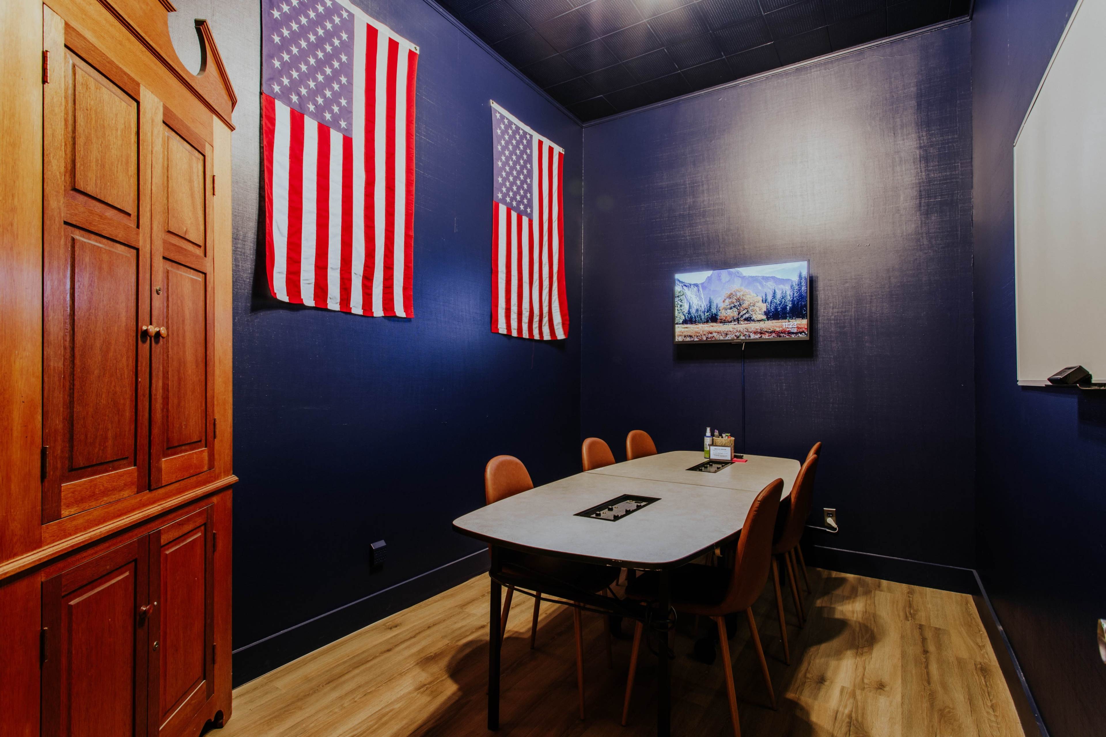 The image shows a conference room with a wooden cabinet, a rectangular table surrounded by six chairs, two American flags on the walls, and a television mounted on one wall.