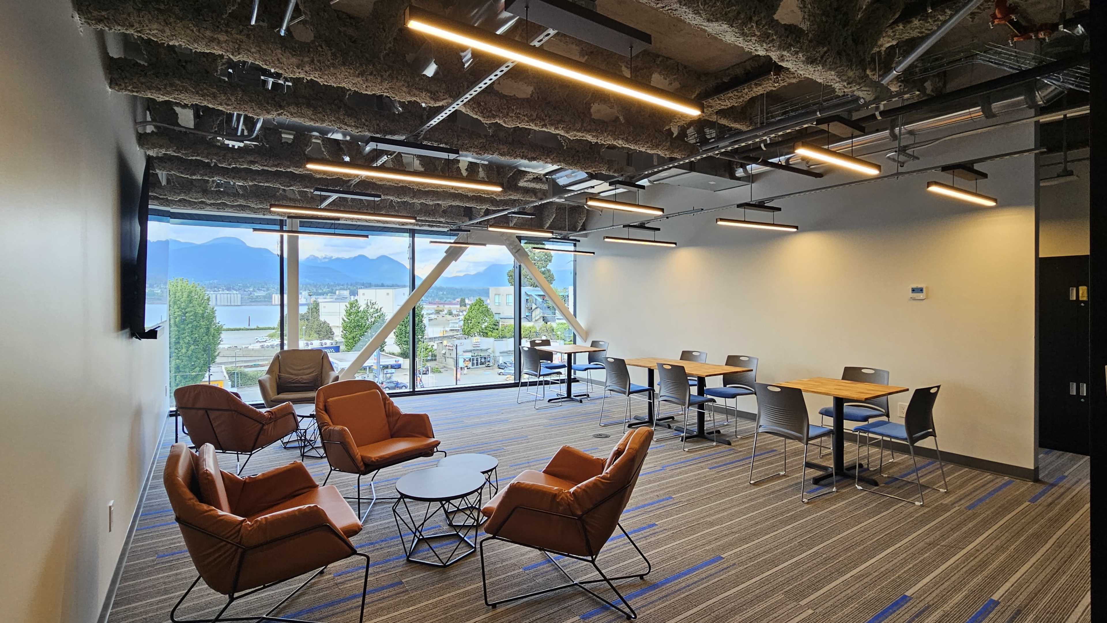 The image shows a modern office space with seating arrangements, large windows providing a view of mountains and water, and tables for working.