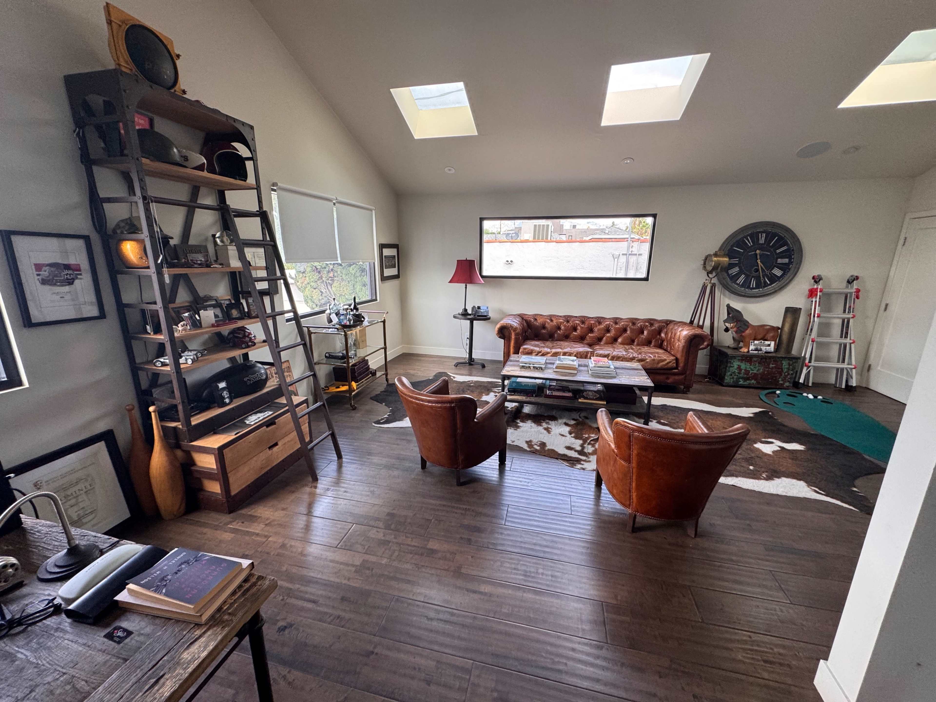 The image shows a modern living room with a leather chesterfield sofa, two brown armchairs, a coffee table stacked with magazines, and a bookshelf against the wall, featuring a mix of decorative items and plants.