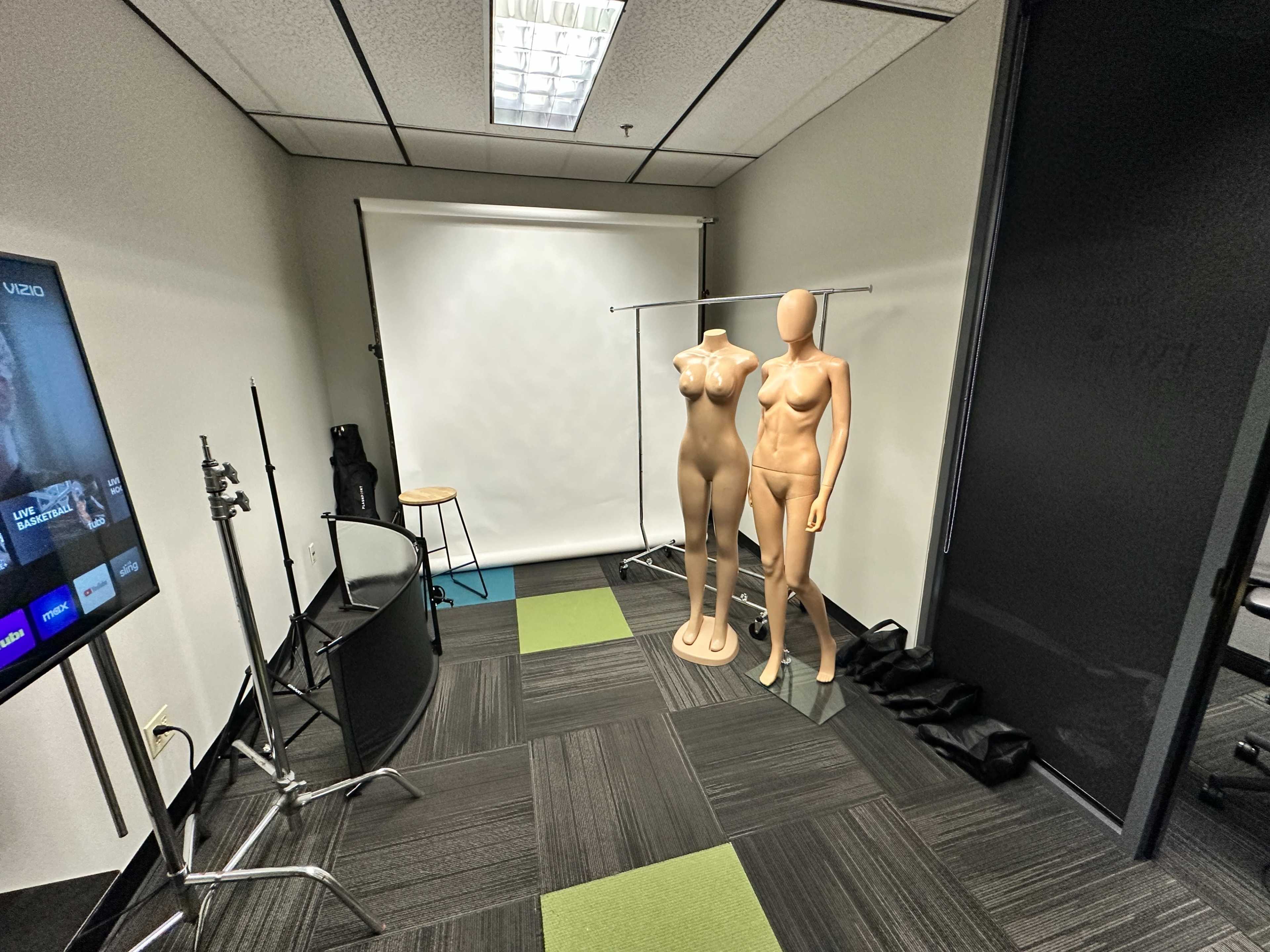 The image shows a small studio space with two female mannequins on display, a black chair, and a TV screen on the wall, along with a backdrop setup.