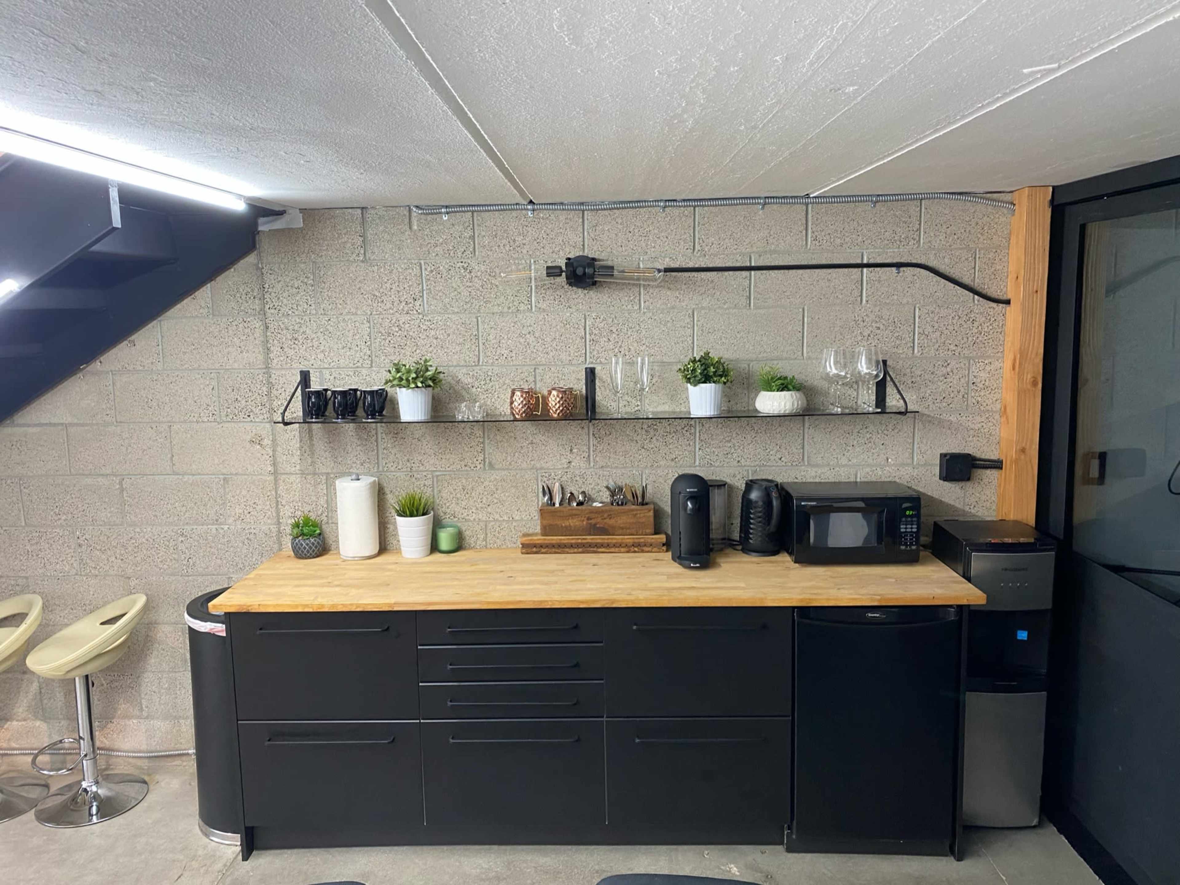 A modern kitchen area featuring a wooden countertop with dark cabinetry, a microwave, a coffee maker, and decorative plants on open shelves against a concrete wall.