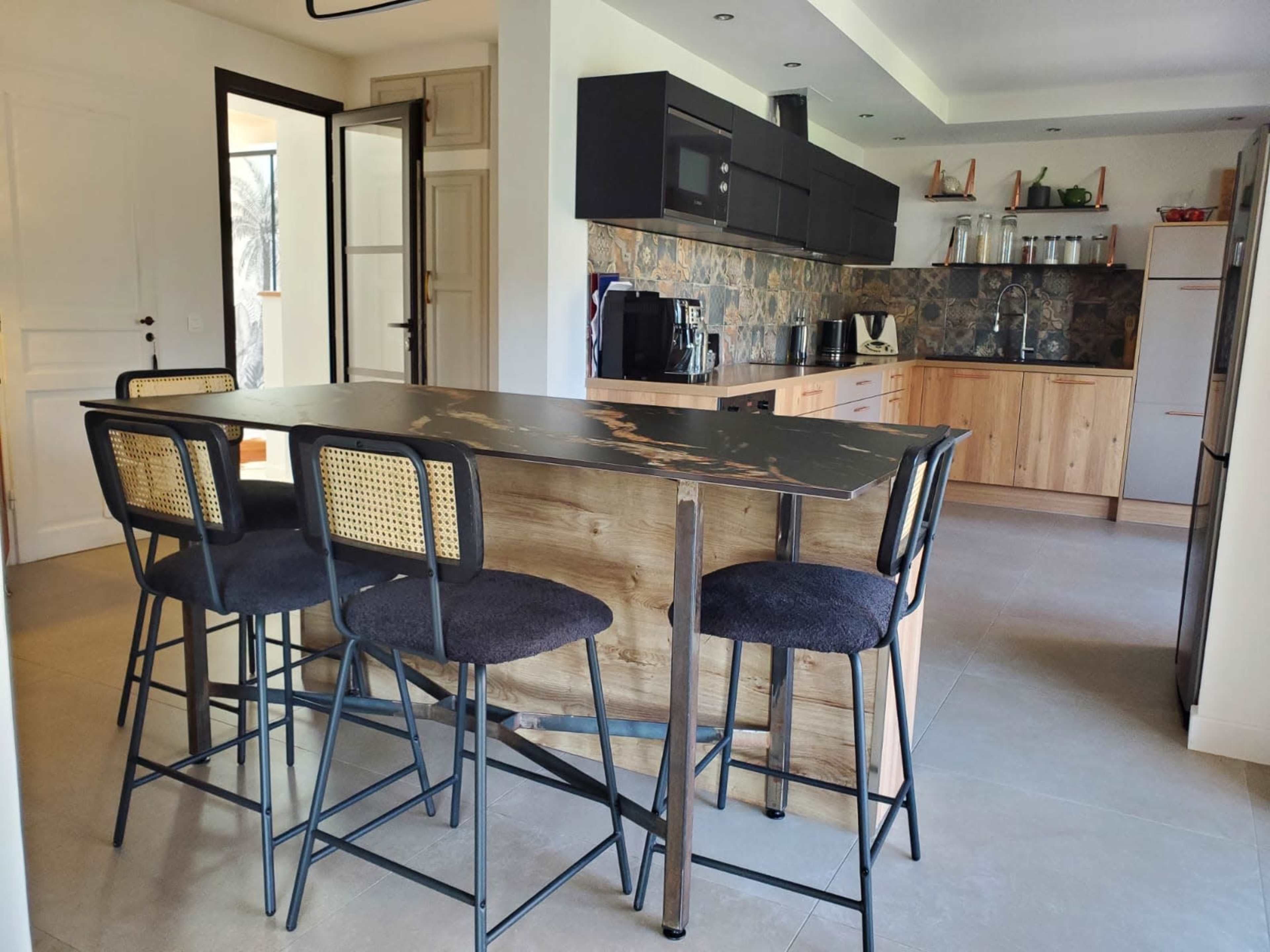 The image shows a modern kitchen with a wooden island bar and four high stools, featuring a coffee machine and various kitchen appliances on the counter.