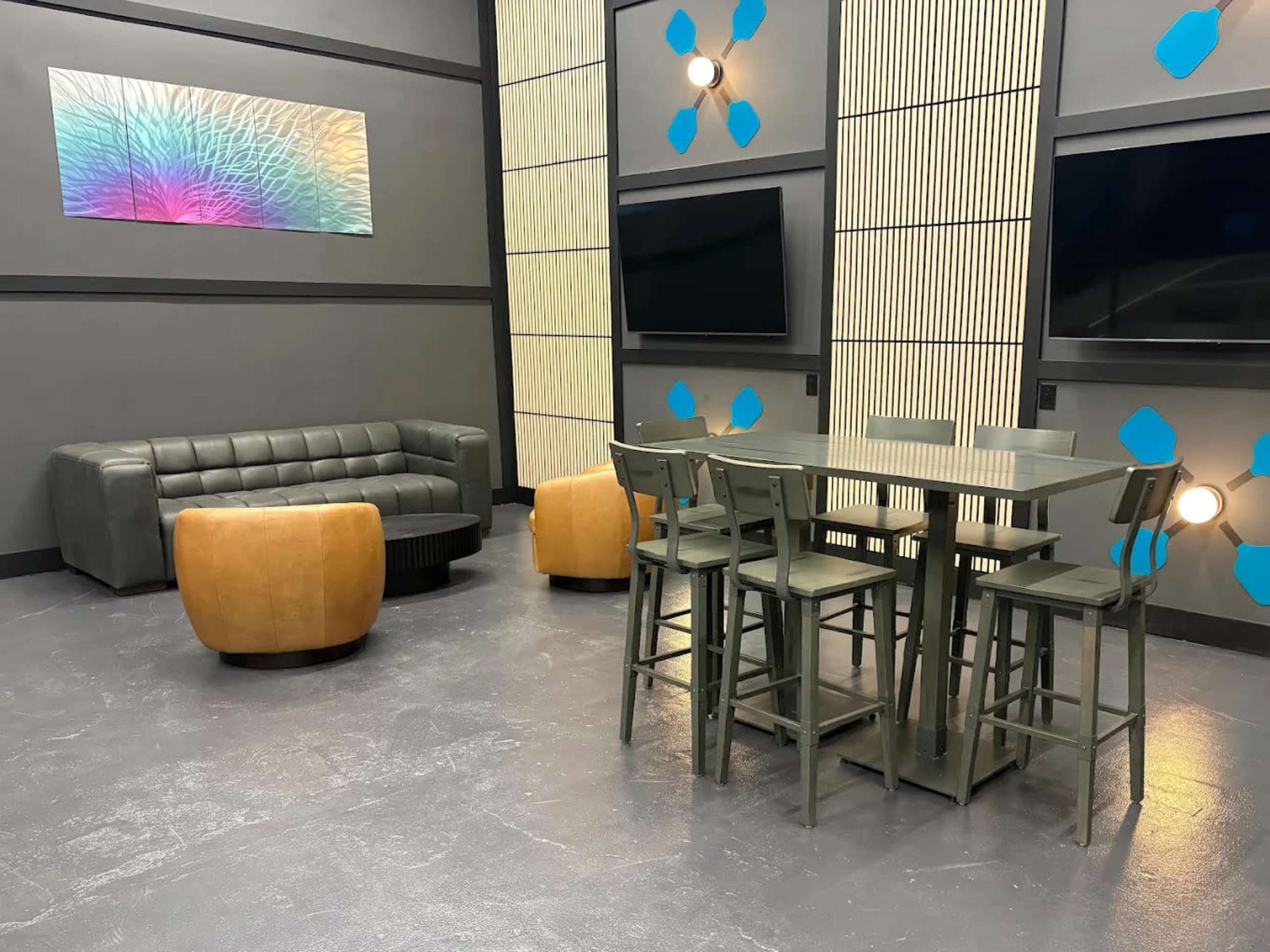 The image shows a modern lounge area with a dark gray floor, a leather couch, two orange accent chairs, a tall dining table with chairs, and several wall-mounted televisions.