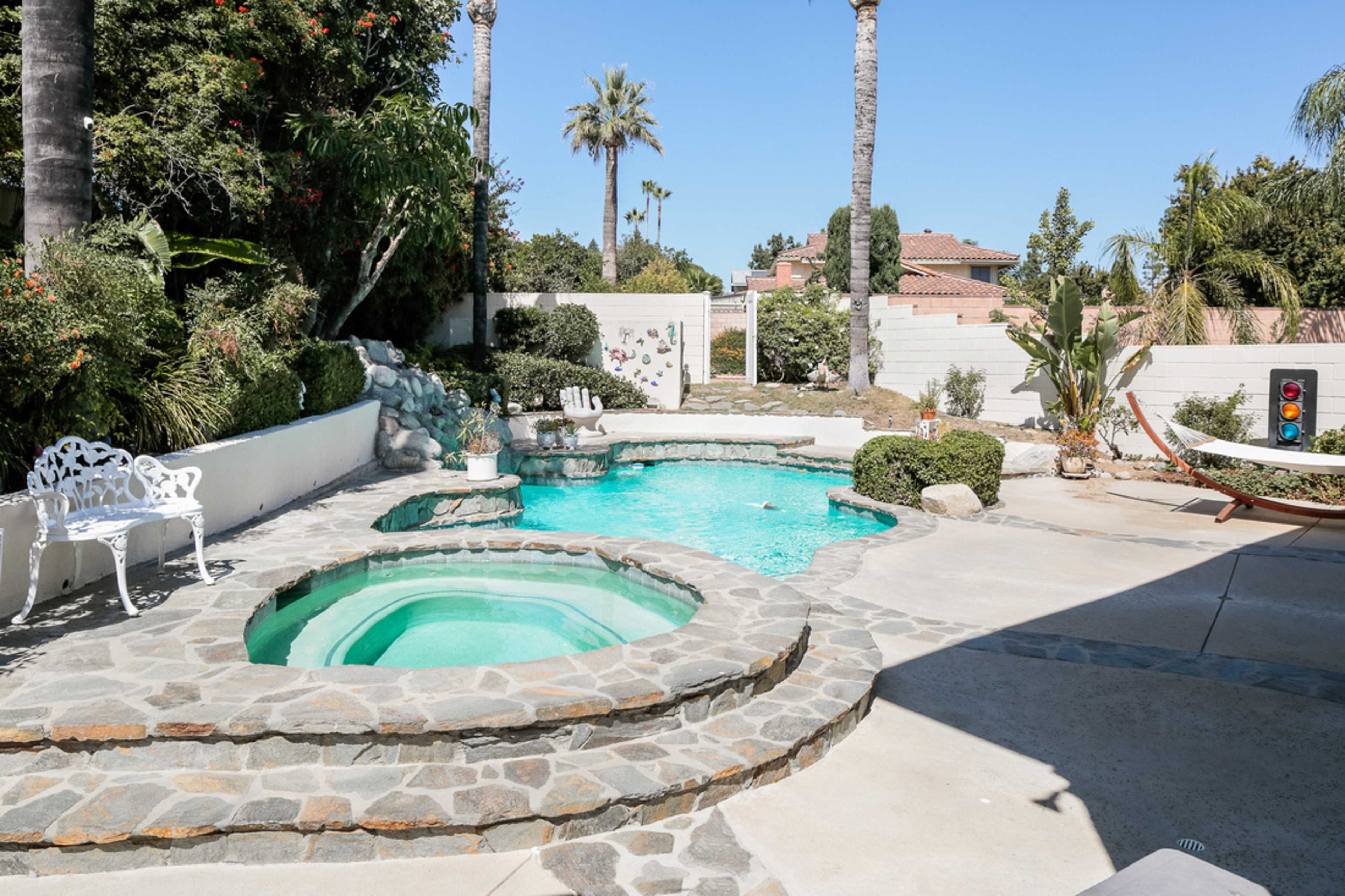 A backyard with a swimming pool and a spa surrounded by stone pathways, palm trees, and landscaped greenery.