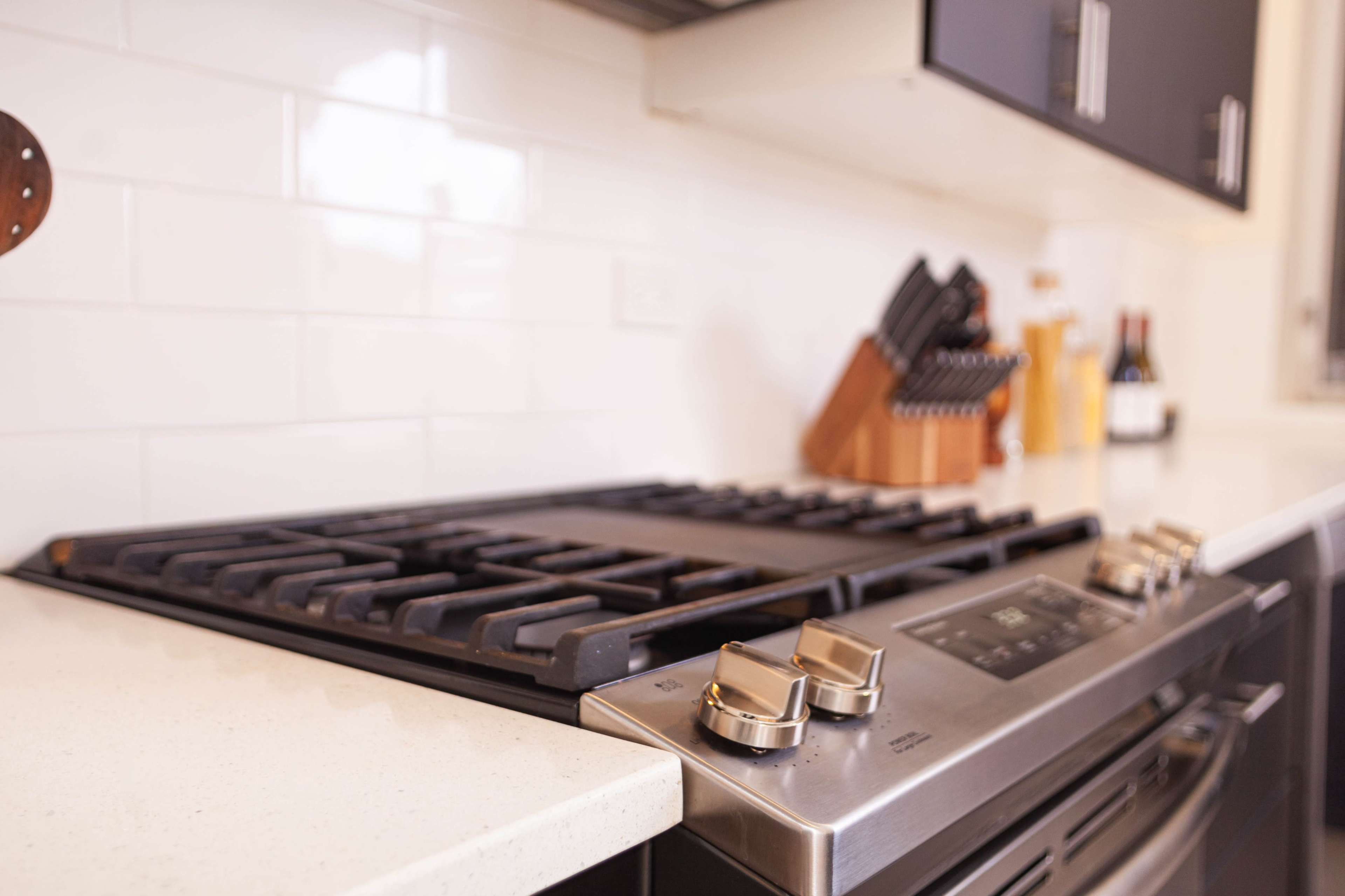 A modern kitchen features a stainless steel stove with gas burners and a granite countertop, accompanied by a knife block and bottles of wine in the background.