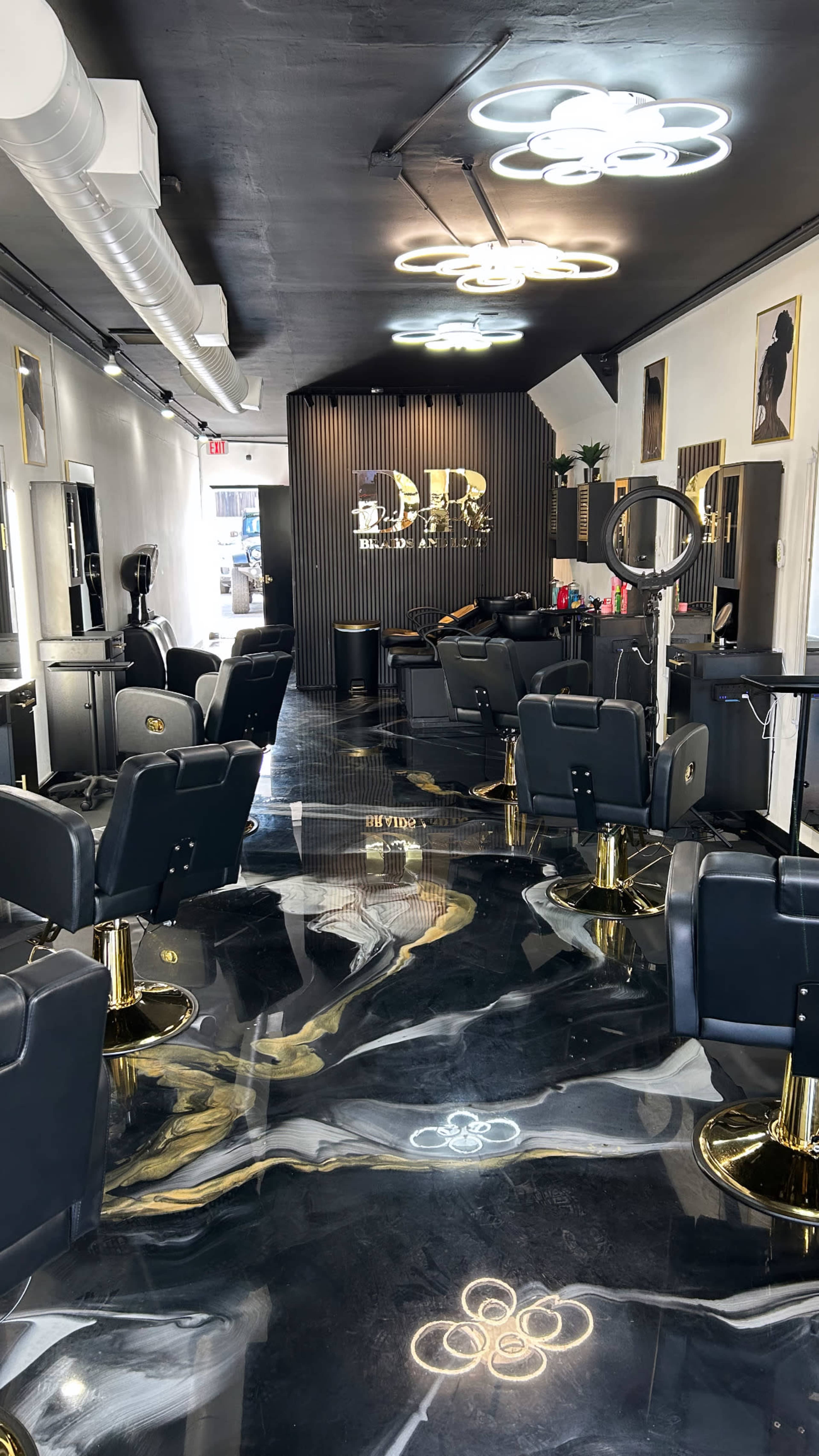 The image shows a modern hair salon with black and gold decor, featuring multiple styling stations, a sleek floor design, and illuminated flower-shaped light fixtures.